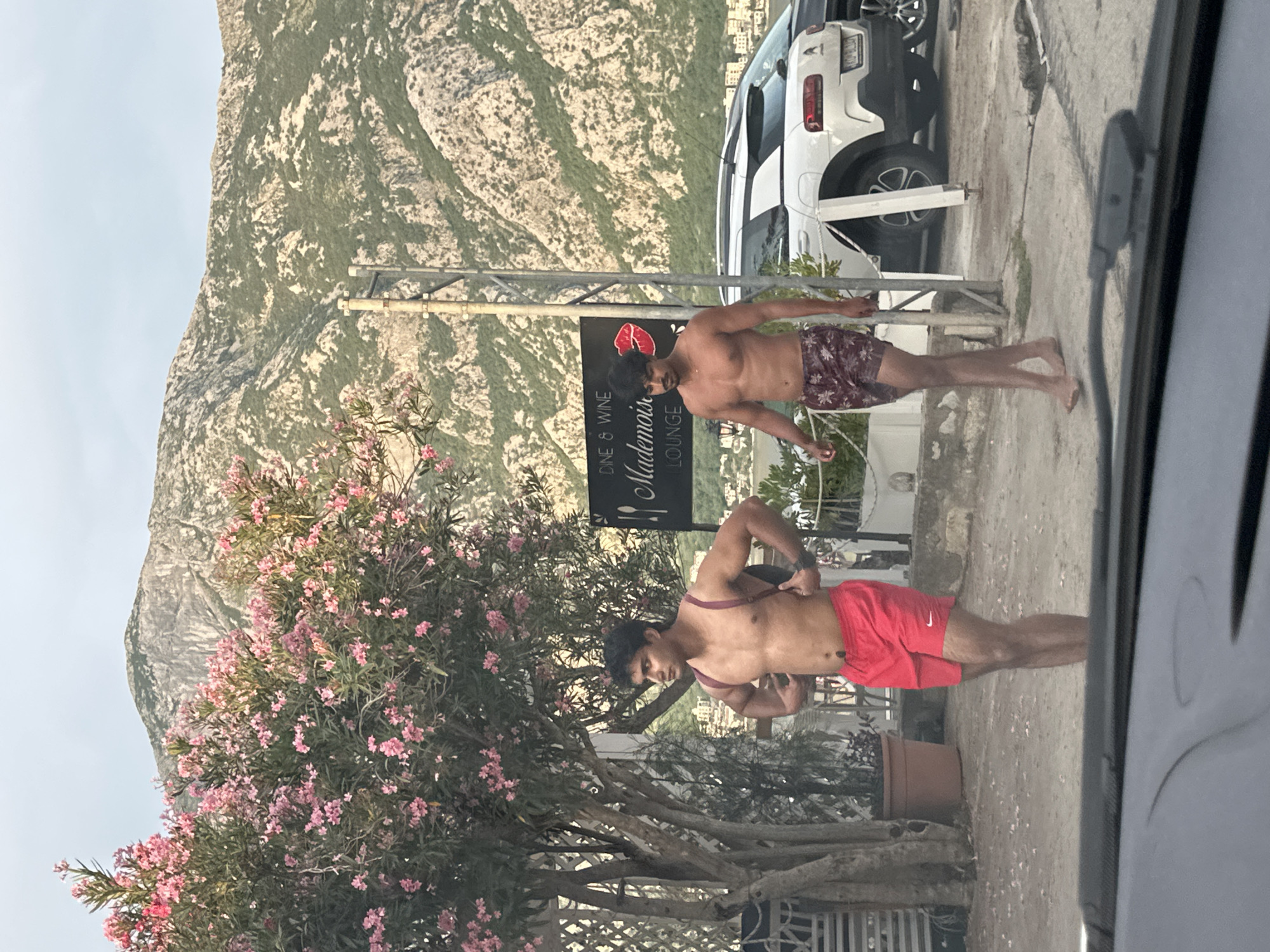 The image shows two shirtless young men standing outdoors. The man in the foreground, on the right, wears red athletic shorts with a white Nike logo. He is adjusting the straps on his shoulders. The man behind him, on the left, wears patterned shorts.
They are standing on a paved area. To their left is a tree with abundant pink flowers. In the background, a large, rocky mountain with sparse vegetation rises behind a white car and a building. A sign with the text "DINE & WINE Mademois Lounge" is visible between the two men.
The scene appears to be outdoors during the daytime, likely with clear weather, given the bright lighting and shadows. The setting suggests a coastal or mountainous region. The overall mood seems casual and relaxed.