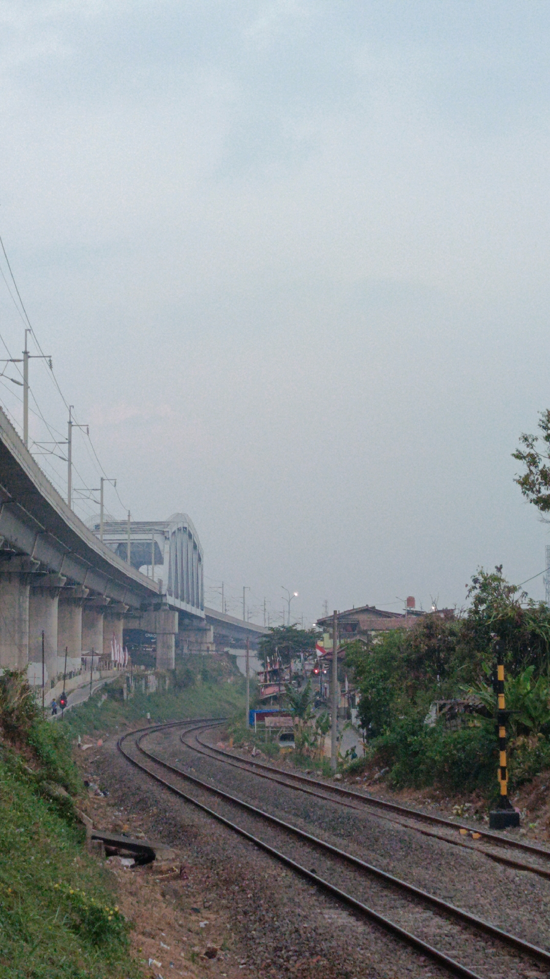 The image depicts a railway line curving through a somewhat overcast urban landscape in Margajaya, Indonesia. In the foreground, a set of parallel train tracks made of gravel and ballast are visible, disappearing into the distance. To the left, a raised concrete railway bridge or viaduct, supported by numerous pillars, runs parallel to the tracks. A smaller, arched railway bridge can be seen further along, partially obscured by the viaduct. Electric power lines stretch across the scene, supported by poles.
On the right side of the tracks, a dense collection of buildings and trees forms a backdrop. Some of the buildings appear to be houses, with visible roofs and windows. A traffic signal with red lights is present, indicating activity on a road that likely intersects the railway. A black and yellow striped signal post stands near the tracks. Lush green vegetation, including trees and bushes, lines the embankment along the railway. A motorcycle is parked on the side of the road beneath the elevated structure. The sky is a pale, hazy grey, suggesting diffused light.