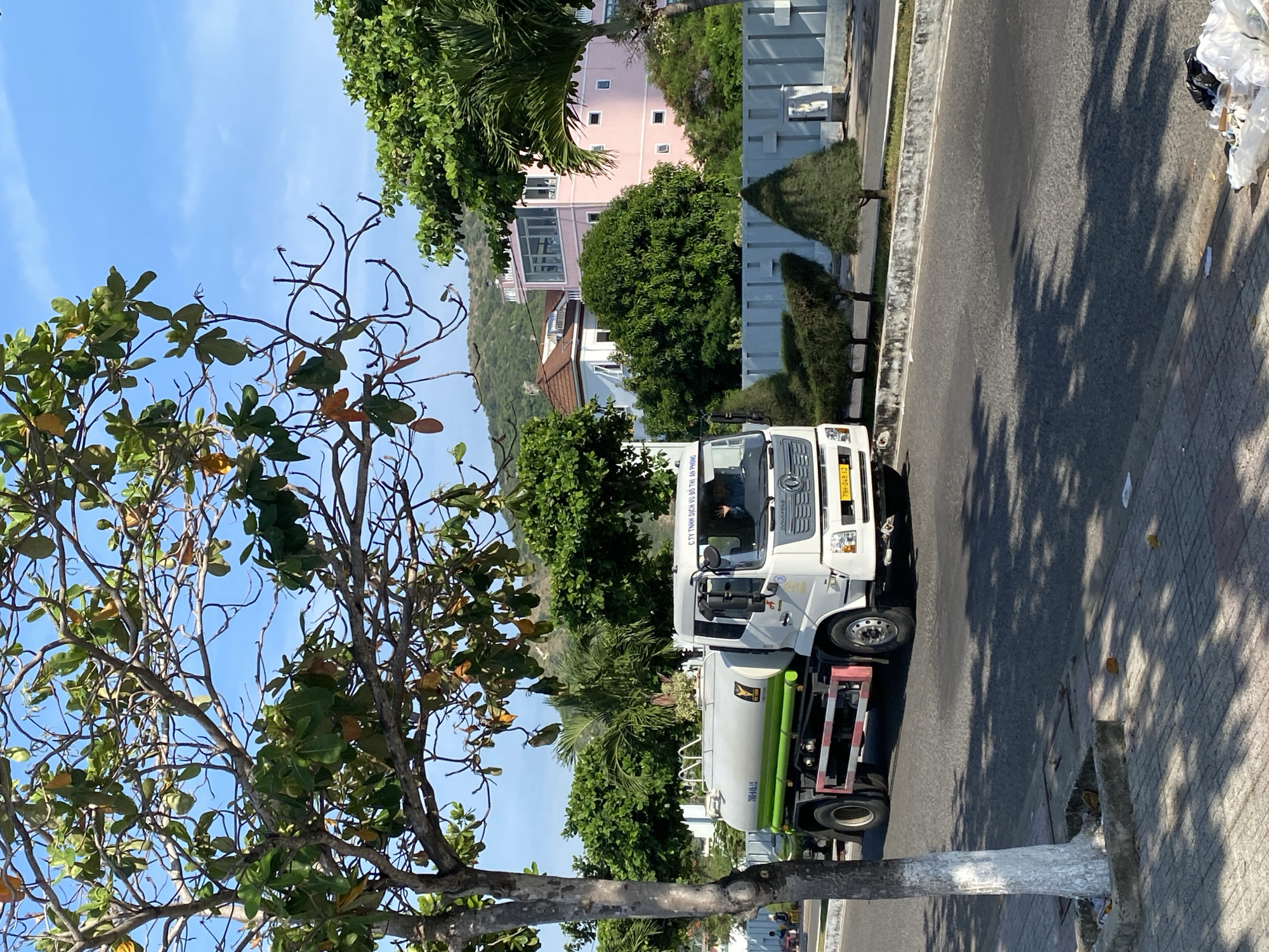 A white water tank truck is driving on a paved road in Bac Nha Trang Ward, Vietnam. The truck has a light green stripe around the tank and is emblazoned with text in Vietnamese. A person is visible in the driver's seat. The road is lined with a sidewalk with paving stones and a tree with white-painted bark. Lush green trees and bushes are present on the left and right sides of the road, with buildings and hills visible in the background. The sky is clear and blue. There is a small pile of trash on the right side of the sidewalk.