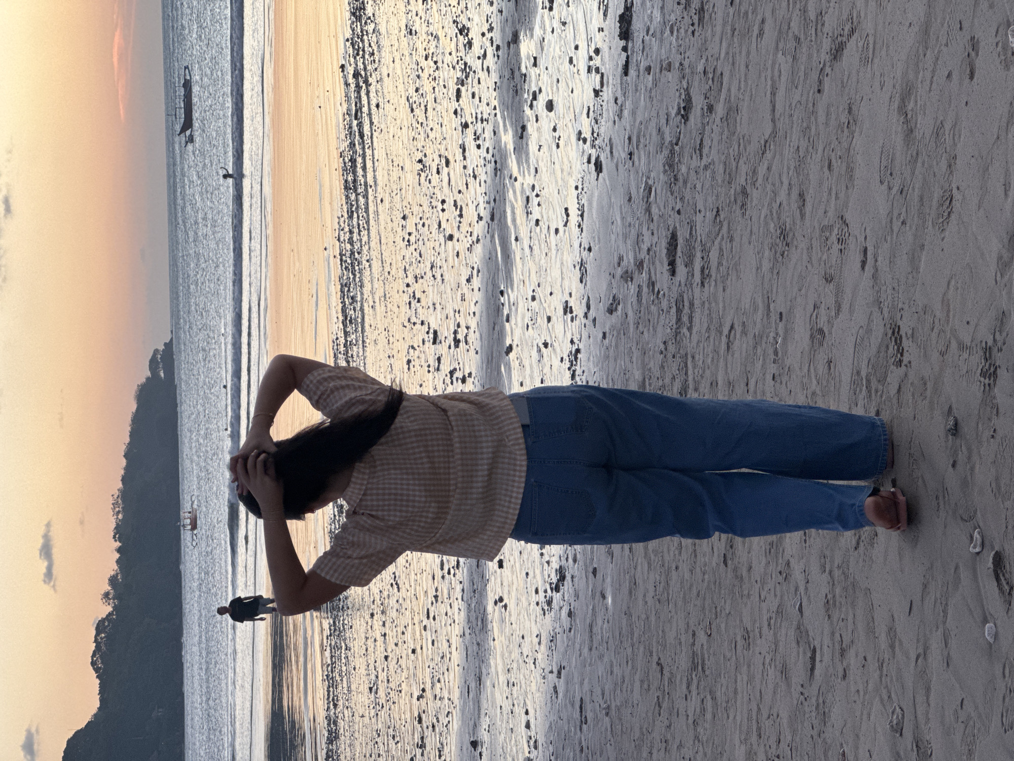 The image displays a serene beach scene during either sunrise or sunset, characterized by warm, soft lighting. The main subject is a person, seen from behind, standing on the sandy shore facing the ocean. This person has long, dark hair which they are holding with both hands at the back of their head. They are wearing a light-colored, possibly checkered, short-sleeved shirt and blue jeans.
In the mid-ground, another silhouetted figure is visible walking on the wet sand closer to the water. Further out on the calm ocean, a traditional boat, possibly a jukung, floats peacefully, with a very faint figure nearby. The sky above is painted with soft orange, pink, and yellow hues, which are reflected on the water's surface. A dark, lush mountain or island forms the backdrop on the left side of the horizon.
The beach itself consists of wet, light-colored sand dotted with dark pebbles and a drier, lighter section with numerous footprints. The overall ambiance suggests a tranquil moment of enjoying the natural beauty of the coastline. The setting is consistent with a tropical beach in Kuta Selatan, Indonesia. There is no visible text in the image.