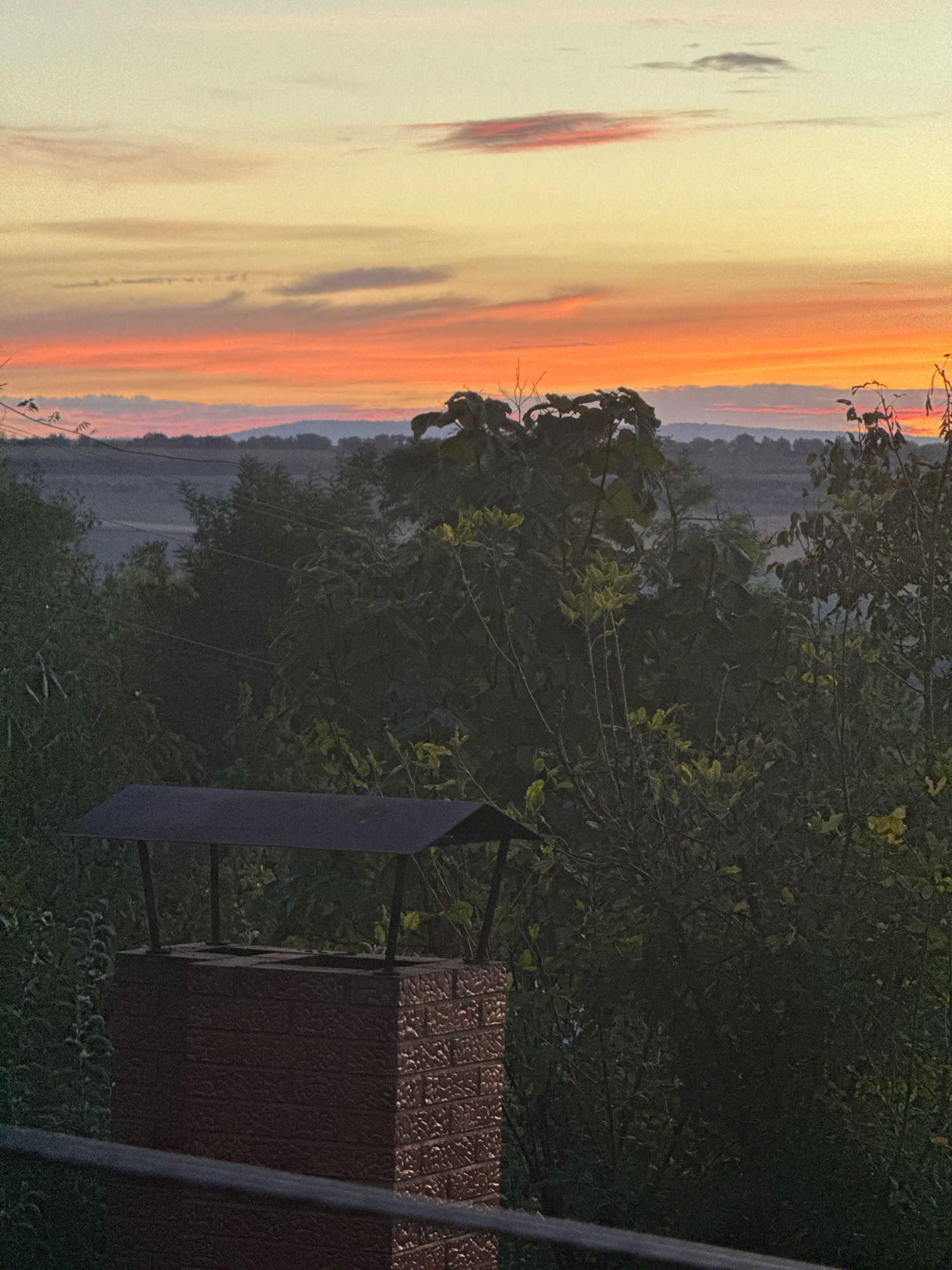 The image captures a sunset over a rural landscape, likely in Elizavetovca, Moldova. In the foreground, a brick chimney with a metal cap stands prominently. Behind the chimney, dense green foliage creates a dark, shadowy screen. Further in the distance, rolling hills are visible, shrouded in a light haze. The sky is a canvas of soft colors, transitioning from a pale yellow at the top to warm oranges and pinks near the horizon, with a few wispy clouds scattered across it. The overall impression is one of peaceful twilight in a natural setting.