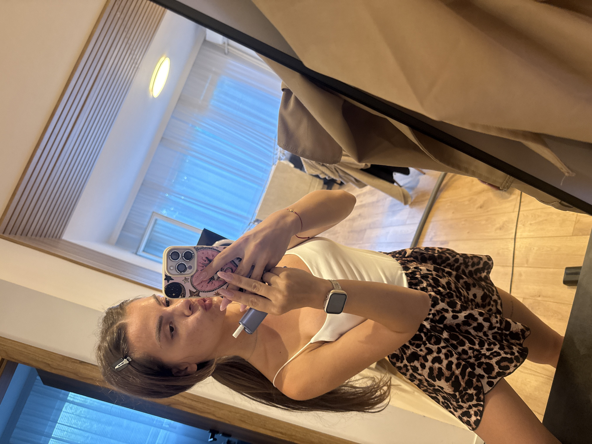 A young woman is taking a selfie in a mirror. She has long brown hair tied in a ponytail and is wearing a white camisole and leopard print shorts. She is holding a blue device in her left hand and her phone in her right hand, which is angled to capture the reflection. She is wearing a watch on her left wrist and a red bracelet on her right arm. The background shows a room with wooden floors, a window with blue curtains, and a light fixture on the ceiling. The image appears to be taken indoors. There is no information available to determine the location in the city of Plovdiv, Bulgaria.