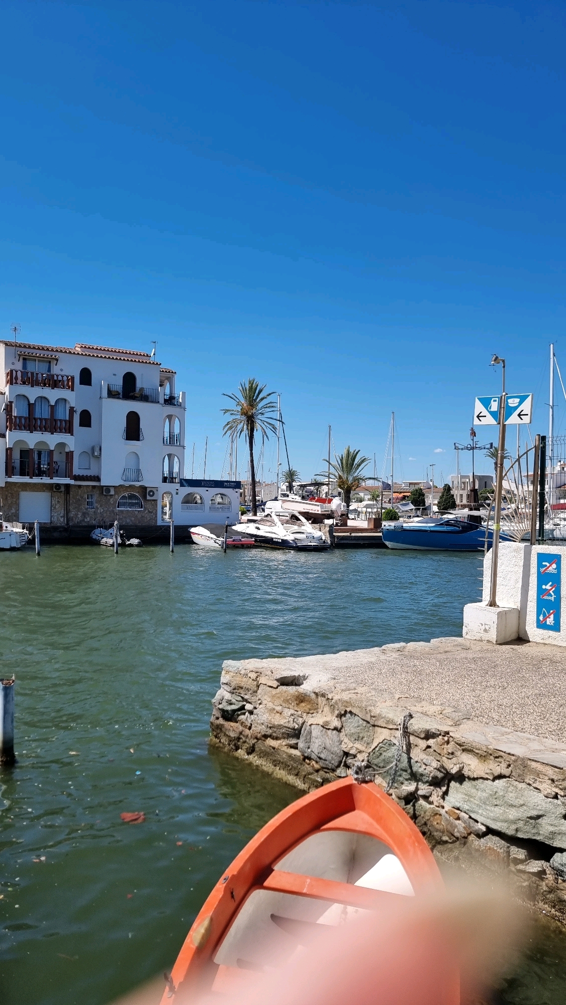 The image depicts a sunny day in the marina of Empuriabrava, Castelló d'Empúries, Spain. In the foreground, the edge of a small orange boat is visible, partially out of focus. The water of the marina is a choppy, greenish hue, with a few pieces of debris floating on the surface. To the left, a white building with multiple balconies and red-tiled roofs lines the waterfront, possibly an apartment complex or hotel. Several boats are docked in the marina, including a white motorboat and a sleek blue yacht. Palm trees are scattered throughout the scene, adding to the Mediterranean atmosphere. In the background, more sailboats and buildings can be seen, along with a clear blue sky. Signs with directional arrows and symbols are posted on a white pillar on the right side of the image. The overall scene suggests a peaceful and scenic vacation destination.