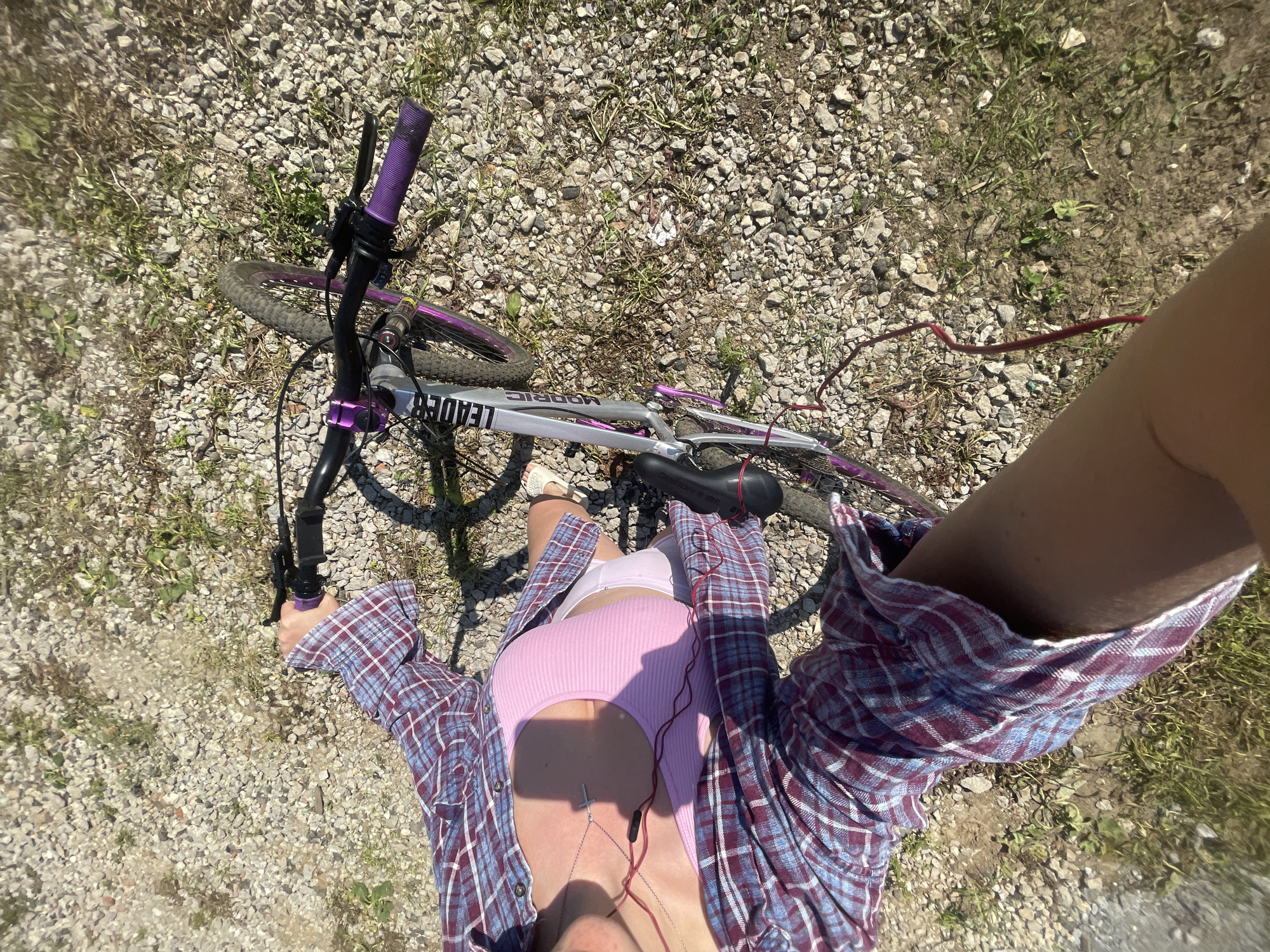 A high-angle, top-down shot captures a person and their bicycle on a sunny day. The person, likely female, is partially visible, dressed in a pink ribbed tank top, a blue and red plaid long-sleeved shirt worn open, and white shorts. A silver cross necklace hangs from her neck, and red wired earphones are visible around her torso. Her right hand grips the purple handle of the bicycle, while her left arm and leg are also visible.
The bicycle, primarily silver, features distinct purple accents on its handlebars and possibly its wheels. The brand names "LEADER" and "MODRIC" are clearly visible on its frame. The bicycle seat, black with a red elastic band, is also in view.
The setting appears to be an unpaved outdoor area, characterized by a ground covered in light-colored gravel mixed with patches of dry grass and dirt. The bright sunlight suggests it is daytime, casting shadows from the bicycle and the person onto the ground. The scene conveys a casual moment, possibly a break during a bike ride. The location context provided is Мосметаллоконструкция Онт, Russia, which suggests an industrial or semi-rural outdoor environment.
