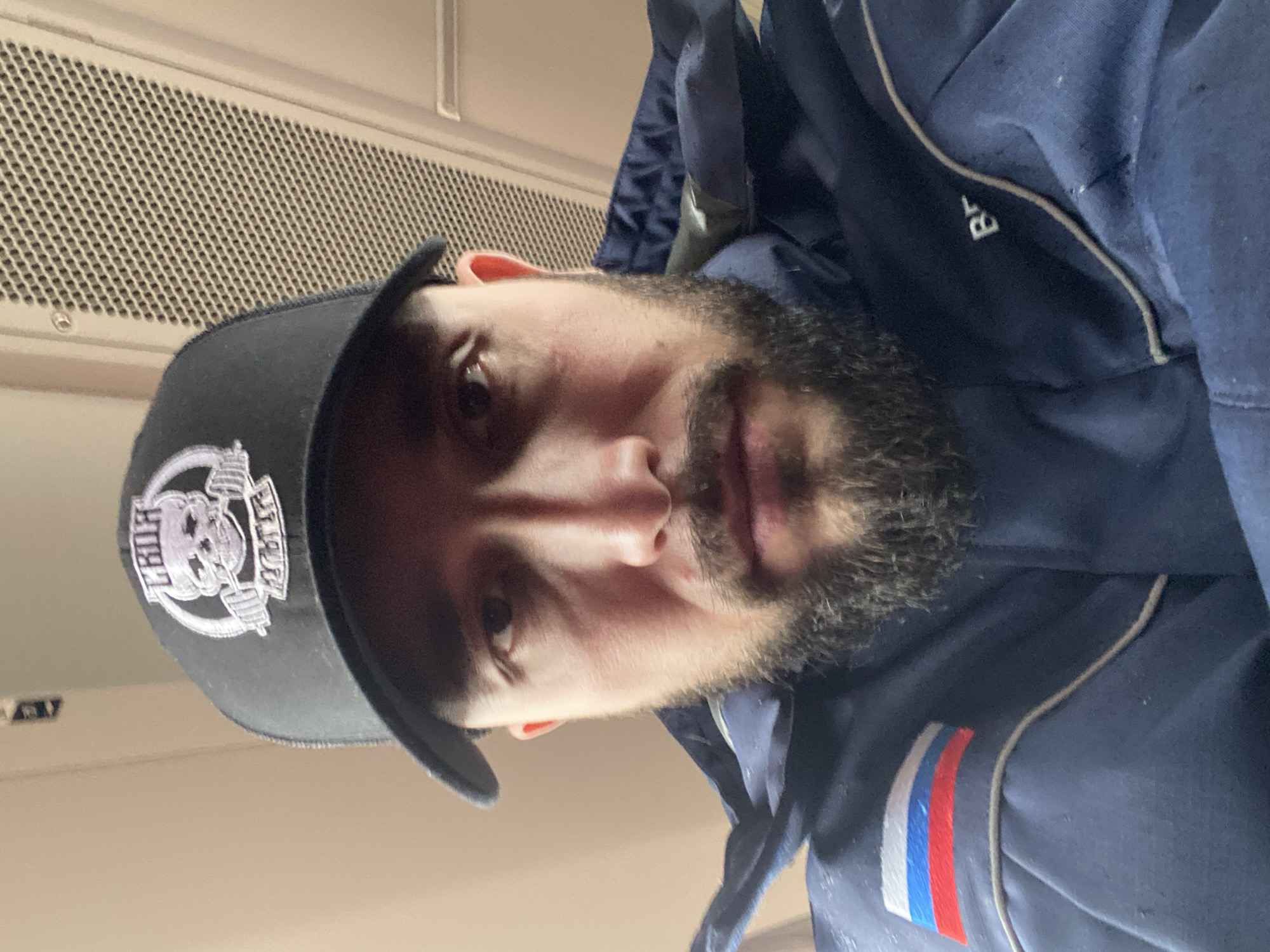 A man with a beard is wearing a black cap with a skull and crossbones logo and a blue jacket with a Russian flag patch on the sleeve. He appears to be inside a building, with a ventilation grille visible in the background. Location information is not determinable based on image.