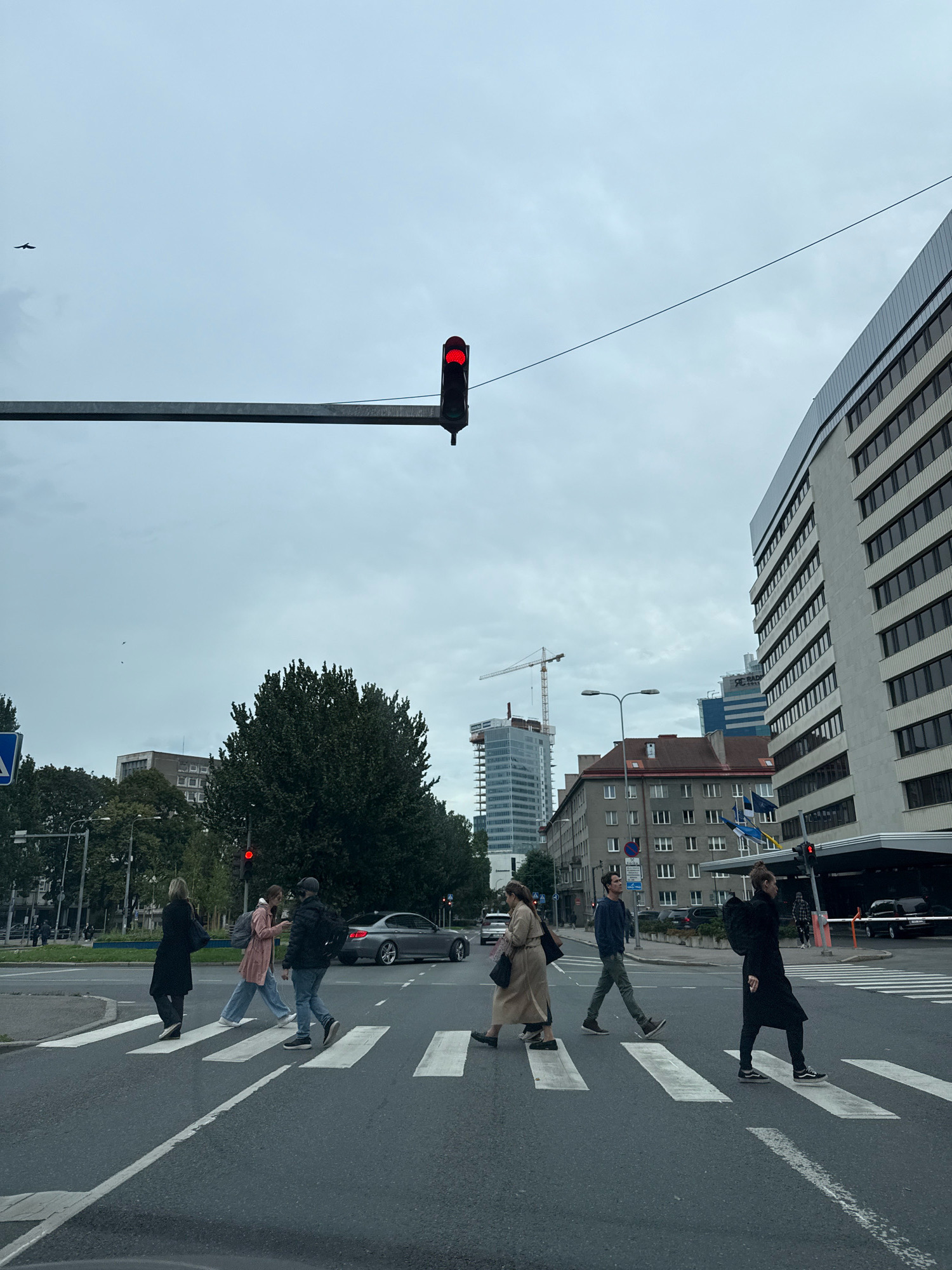In Tallinn, Estonia, on an overcast day, a group of pedestrians is crossing a wide, multi-lane intersection at a marked crosswalk. The traffic light visible above the crosswalk is red, indicating that vehicles must stop. Several people are mid-stride across the zebra stripes, dressed in casual outdoor clothing. A modern, multi-story building with a grey facade and numerous windows dominates the right side of the frame. In the background, more buildings, some appearing to be under construction with cranes, line the city streets, along with trees and streetlights. The sky is a pale grey, suggesting a cloudy or potentially rainy day. A dark-colored car is stopped on the road in the mid-ground, to the right of the pedestrians.
