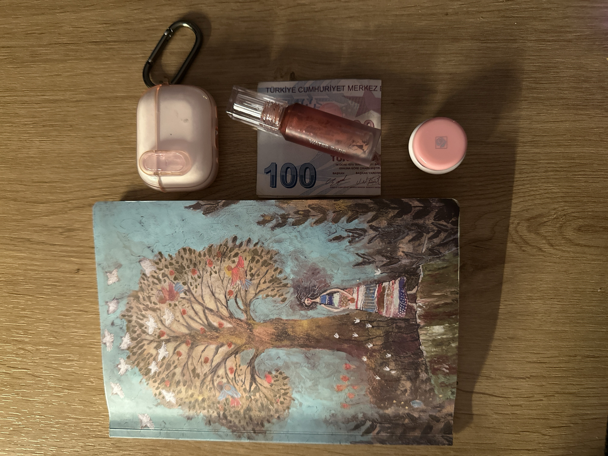 The image displays a collection of personal items arranged on a wooden surface. To the left, a notebook with an artistic cover featuring a tree, birds, and a woman is visible. To the right, a light pink earbud case with a black carabiner clip is positioned. Below the earbud case, a Turkish 100 Lira banknote lies flat, with a small, reddish liquid cosmetic product on top of it. Further down, a small, round, light pink container with a white logo is placed. The lighting suggests an indoor setting, possibly a desk or table, with a shadow cast from the objects. There is no discernible location in the city of Fethiye, Turkey, or any indication of urban or natural environment beyond the immediate foreground. No individuals are present in the scene, and no actions or interactions are depicted.
