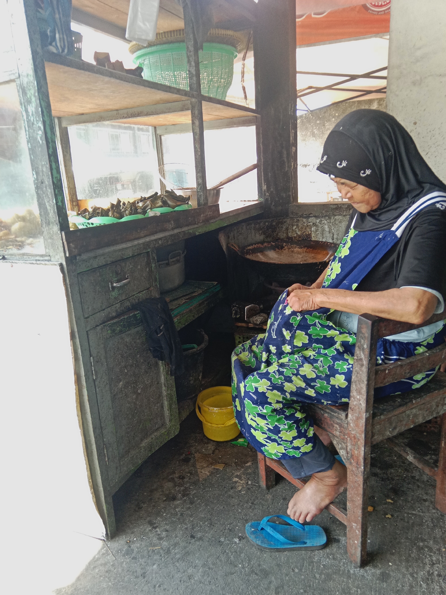 An elderly woman wearing a black hijab and a blue apron with a green clover pattern sits on a wooden chair in front of a food stall in Ringinanom, Indonesia. She is wearing a black shirt and dark pants, and blue flip-flops on her right foot. Her hands are clasped in her lap. The food stall has a weathered green wooden cabinet with a metal handle and a yellow bucket on the floor next to it. On a shelf above the cabinet are various food items, some of which appear to be wrapped in leaves and placed in green baskets. Behind the woman, a large metal pan is visible, suggesting that food is being prepared or cooked. The environment appears to be outdoors, with sunlight casting shadows on the ground. The stall itself is made of wood and possibly metal, showing signs of age and use.