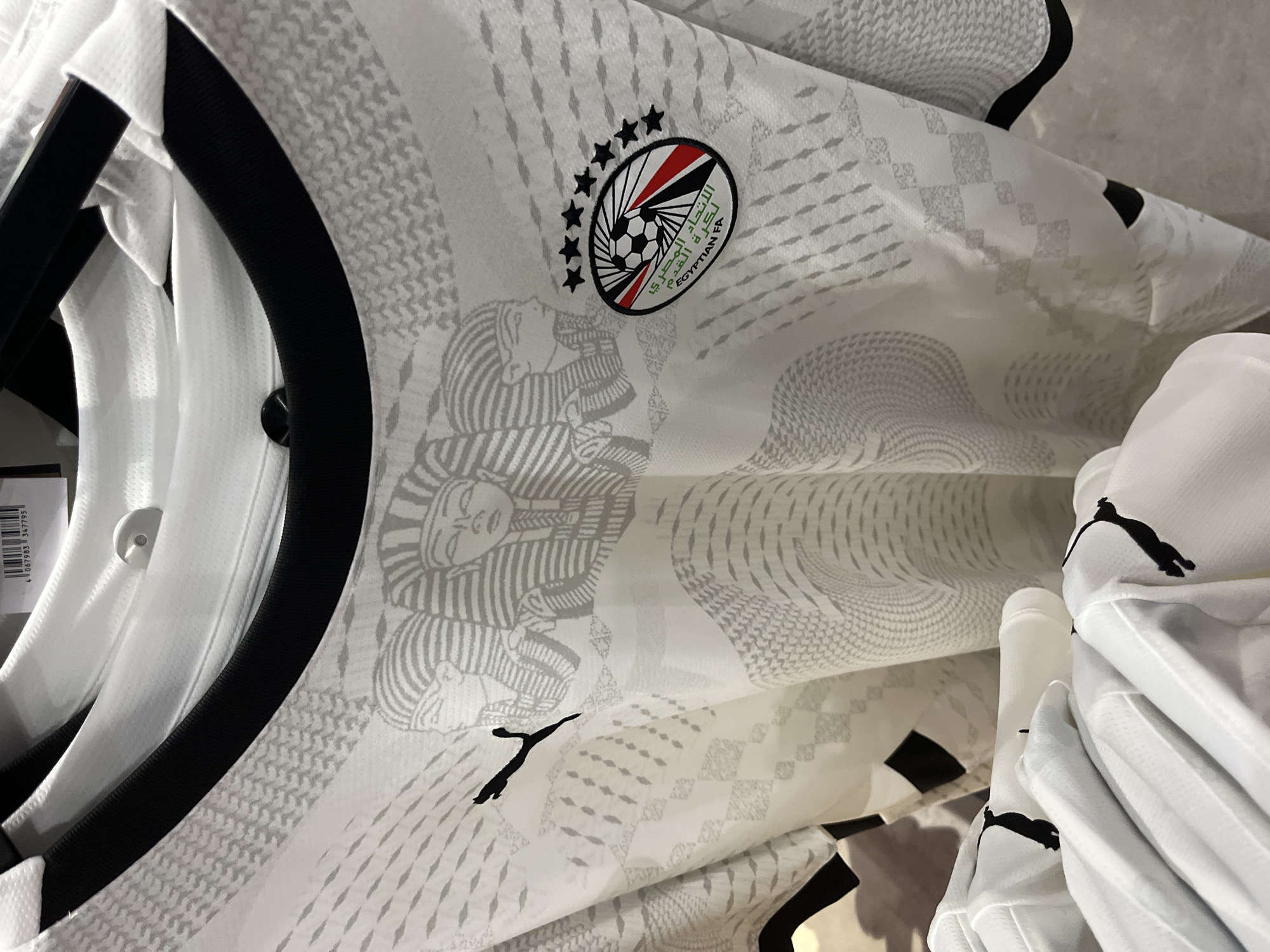 The image is a close-up, eye-level shot of a white football jersey with a black collar. The jersey features a subtle, repeating pattern in a lighter shade of gray, incorporating geometric shapes and what appear to be stylized Egyptian pharaoh heads. On the left side of the chest, a black Puma logo is visible. On the right side of the chest, an oval-shaped emblem is present, containing a soccer ball, stars, and Arabic text which translates to "Egyptian FA". The jersey is hanging, and part of another white jersey is visible in the foreground, also bearing a black Puma logo on its sleeve. The background is out of focus but suggests a retail environment, possibly a sports apparel store, with clothing racks and other garments. There are no people or specific landmarks indicating a location in Rodengo, Italy. The lighting is even, illuminating the details of the jersey.
