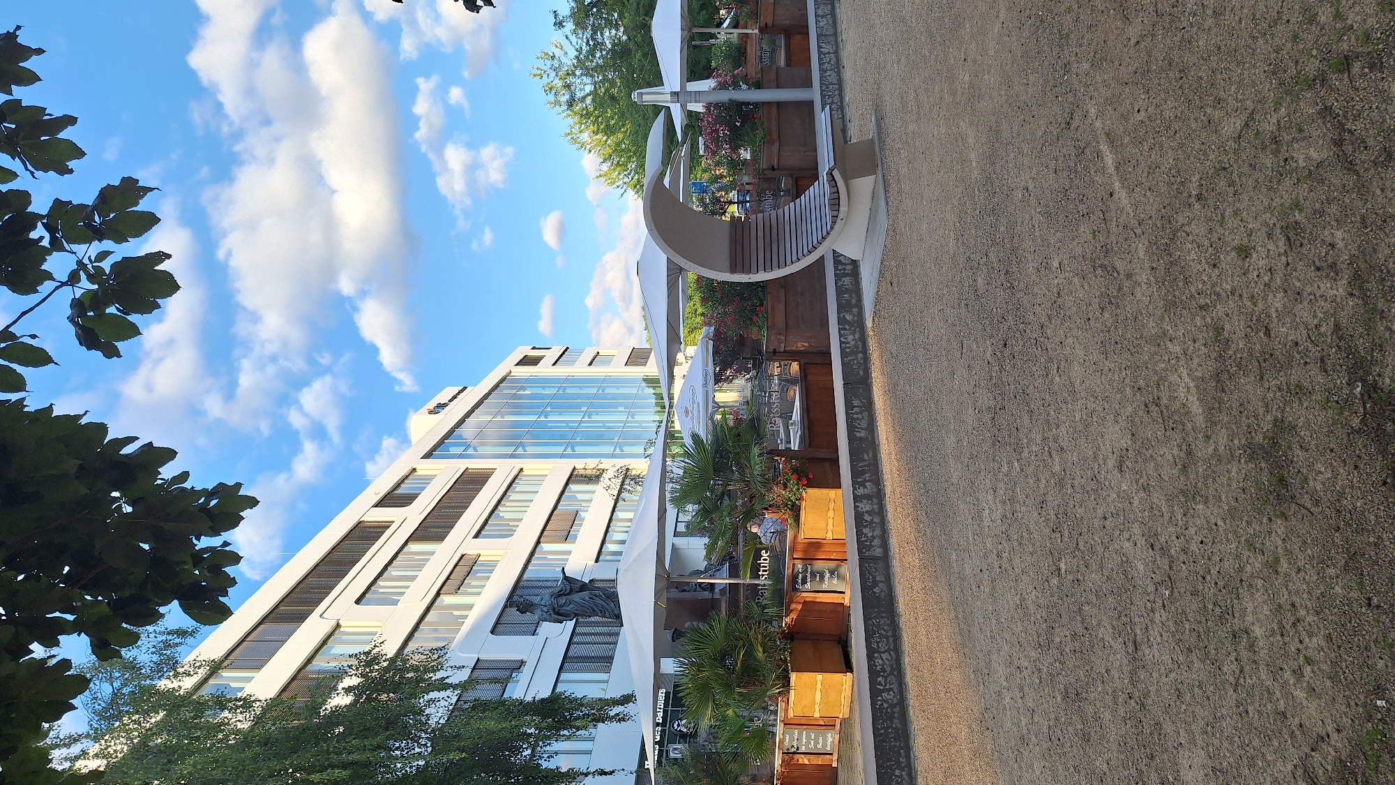 This image depicts an outdoor seating area in front of a modern, multi-story white office building. The building has a prominent glass facade on its lower levels and numerous windows on the upper floors. In front of the building, there are several large white umbrellas providing shade over tables and chairs, suggesting a restaurant or café. Planters filled with palm trees and flowering plants are arranged along the seating area. A statue of a robed figure stands near the entrance to the seating area. To the right, a unique, crescent-shaped modern bench made of concrete and wood is visible. The ground in the foreground is covered in gravel. The sky is blue with scattered white clouds. The image captures an urban scene, likely during daytime, with the office building and outdoor seating area forming the primary subjects. The location is identified as Chemnitz, Germany.