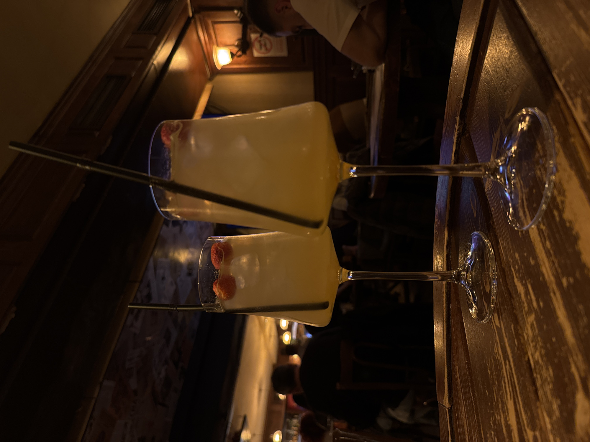 The image shows two cocktails, likely alcoholic drinks, in what appears to be a bar or restaurant in an unknown city. The drinks are in stemmed glasses and have a pale yellow color. Each glass has two red berries (possibly strawberries or raspberries) as garnish inside the glass. A black straw is visible in each glass.
The glasses are on a dark, wooden table. The background is dimly lit, with elements of the bar visible, including wooden walls, hanging lamps, and other people. One person is partly visible at the top of the image, wearing a white t-shirt. Additional people are partially visible towards the bottom of the image.