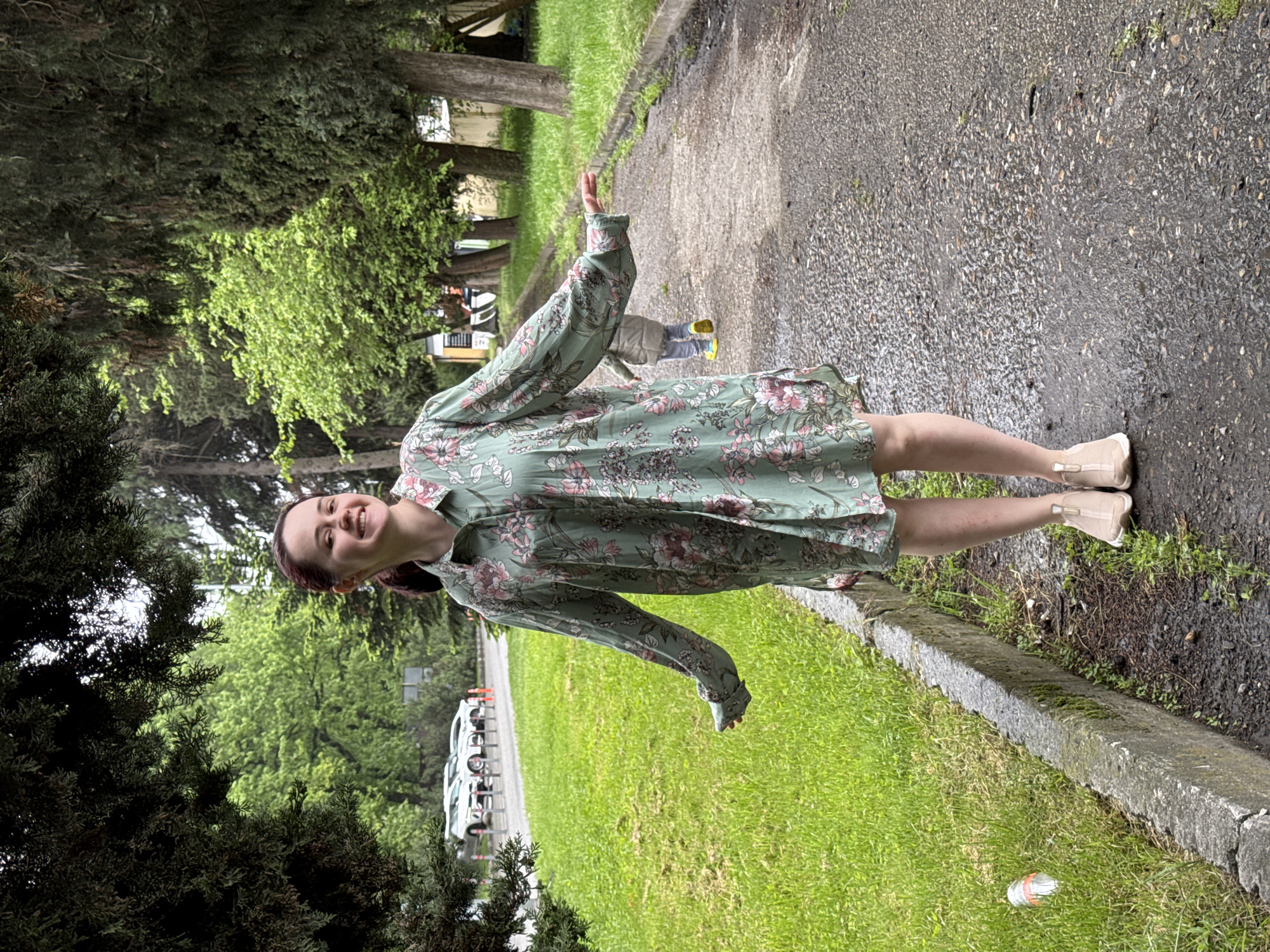 A woman stands on a narrow asphalt path in Sochi, Russia. She wears a light green dress with a floral pattern, tan shoes, and is smiling. The path borders a grassy area on one side and a concrete curb and trees on the other. In the background, cars are parked along the road. A child is partially visible in the distance.