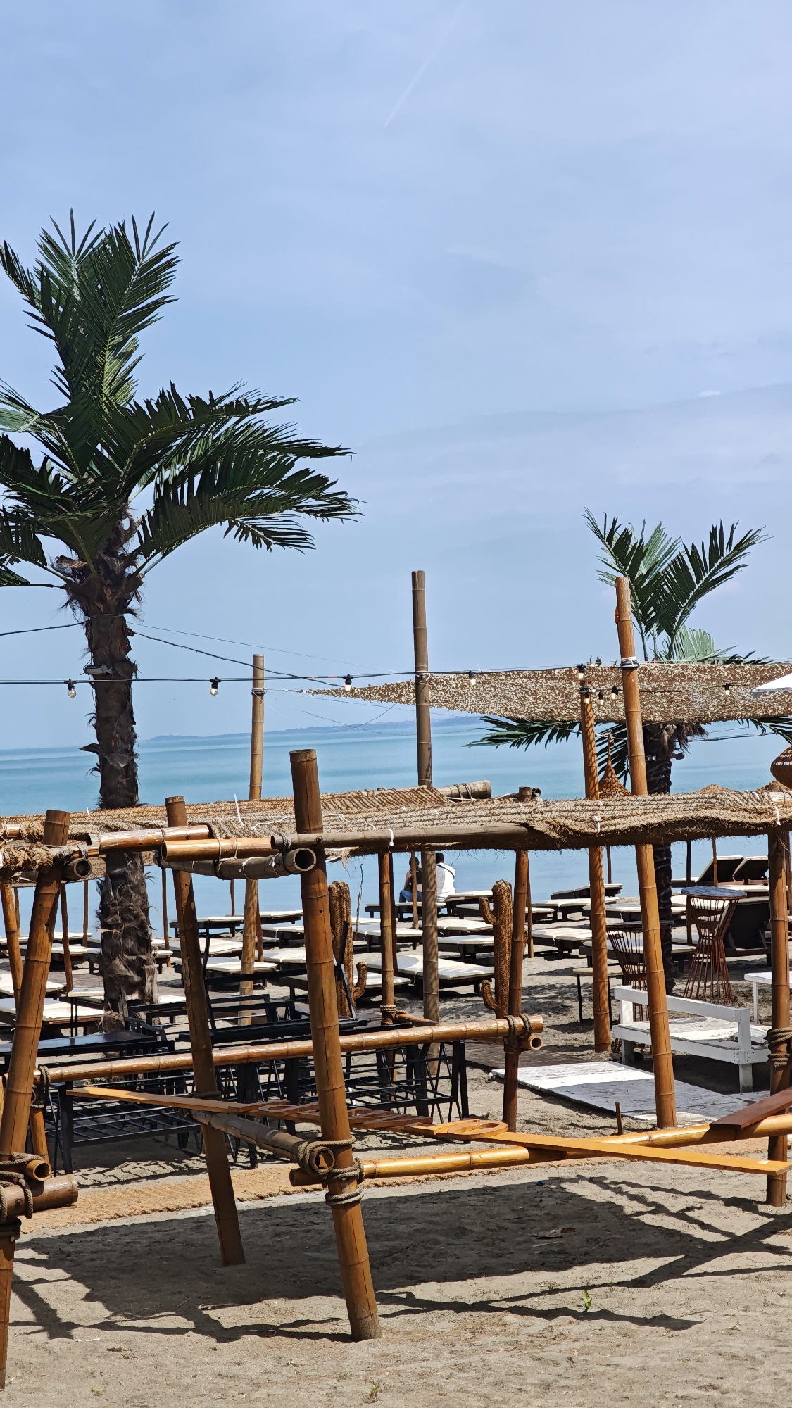 The image depicts a beach scene in Burgas, Bulgaria. It features a beachfront establishment with bamboo structures providing shade and support for tables and seating areas. The structures have thatched roofs. Several rows of beach chairs and tables are visible. Two palm trees are in the background. The sea is visible, with a clear, blue sky overhead. There are people in the background, some seated at tables. The overall atmosphere is relaxed and beachy. The foreground is dominated by the bamboo framing of the structures.