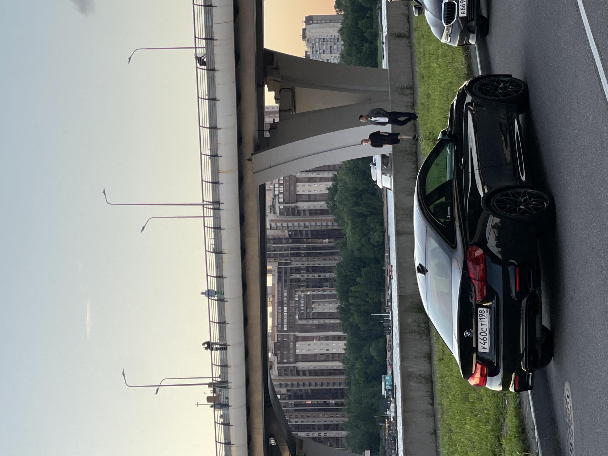 The image shows a city scene with a large bridge in the background, multiple buildings, and a body of water. In the foreground, there are two black cars on a paved road. The car on the left is a coupe, and its license plate is visible, showing "Y460CT 198 RUS". The car on the right is a sedan.
On the bridge, there are several people walking, including a person on a bicycle or scooter. Under the bridge, two men are standing near a concrete barrier. The environment appears to be a park or waterfront area with green grass and trees. The sky is light blue, suggesting it might be late afternoon or early evening.