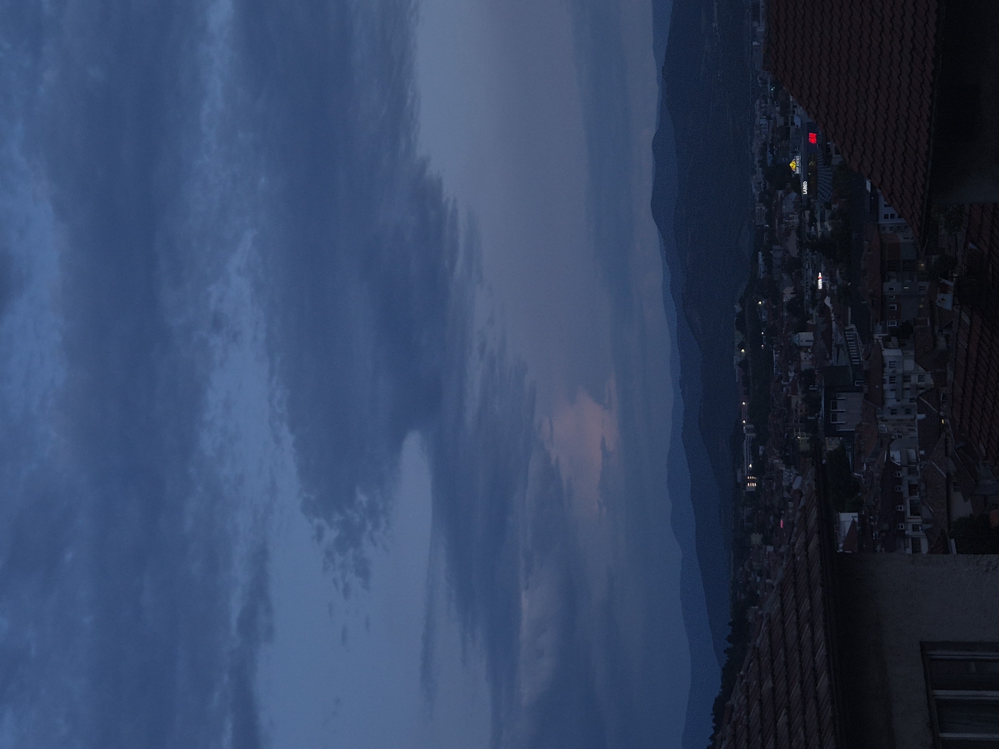 An aerial view captures a cityscape at twilight or night, looking over a densely built area towards distant mountains under a cloudy sky. The image appears to be rotated 90 degrees clockwise from its natural orientation; when viewed upright, the horizon runs horizontally across the middle and right side.
The sky occupies the majority of the frame, showcasing a dramatic expanse of dark blue and grey clouds. Patches of lighter, almost pinkish, light are visible near the horizon, suggesting either the last vestiges of sunset or the first hints of dawn. In the distance, dark, silhouetted mountains form a rugged backdrop against the sky.
Below the mountains, the city stretches out, a mosaic of buildings with numerous red-tiled roofs. Streetlights and building lights illuminate parts of the urban sprawl, casting a warm glow against the deepening darkness. Several illuminated signs are visible: a prominent yellow sign clearly displays the text "HAMBURG MARKET" and "LARGO" below it, while a bright red rectangular sign shows indistinct white letters. Other smaller, unreadable illuminated signs dot the cityscape.
In the bottom right foreground, a dark red-tiled rooftop with a concrete edge provides a vantage point for the scene. Part of a building, including a small, dark window, is visible in the extreme bottom right corner. The overall mood is peaceful and serene, reflecting the quiet transition of time between day and night.