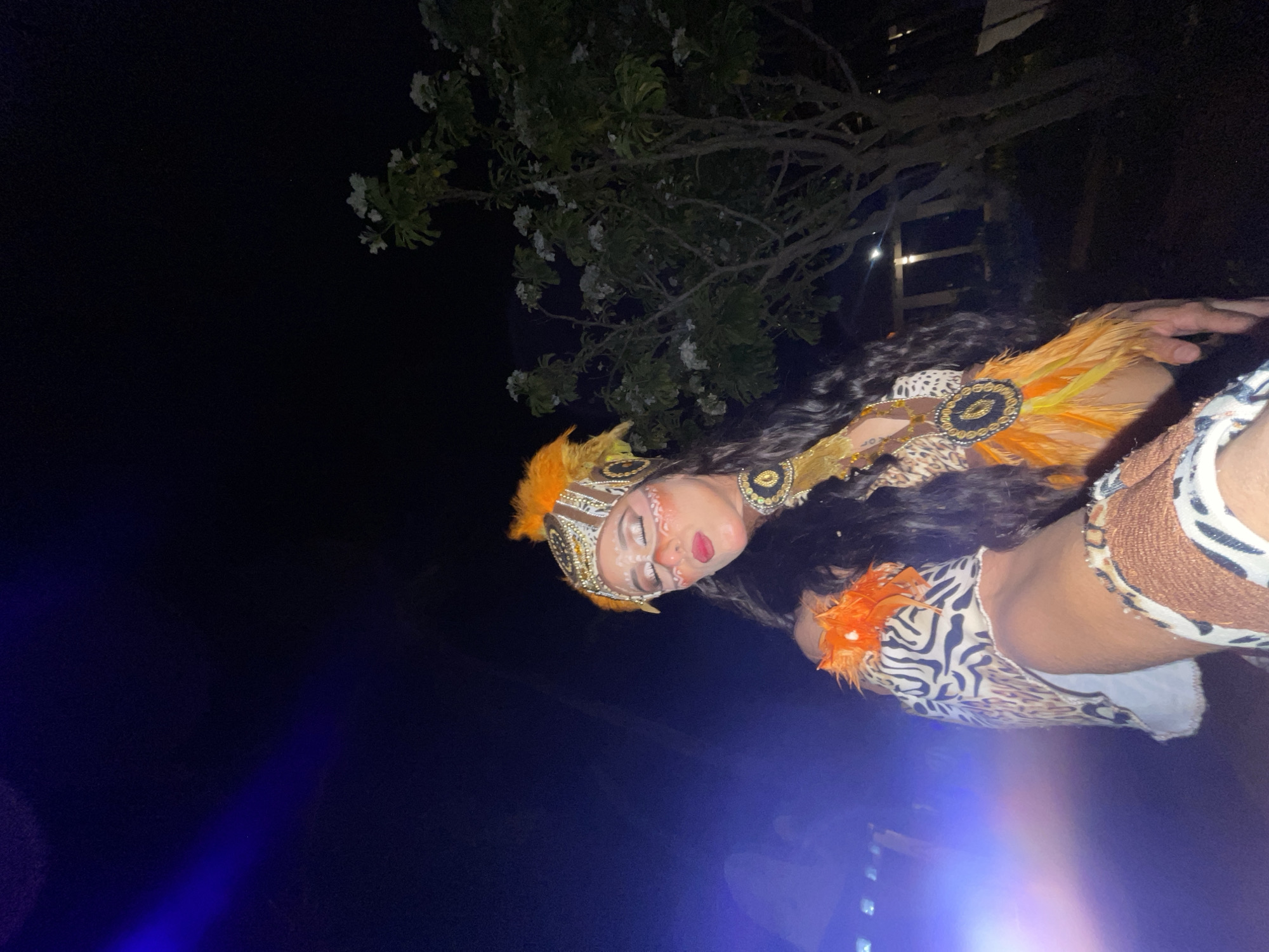 A woman in elaborate indigenous-style costume with face paint and orange feathers is posed against a dark outdoor background at night. She is wearing a headdress, a leopard print garment, and a necklace adorned with feathers. The setting appears to be a garden or outdoor venue, with a tree with white flowers visible to the right, and a blurry blue light on the left, possibly from stage lighting. The scene is likely in Porlamar, Venezuela, given the context.
