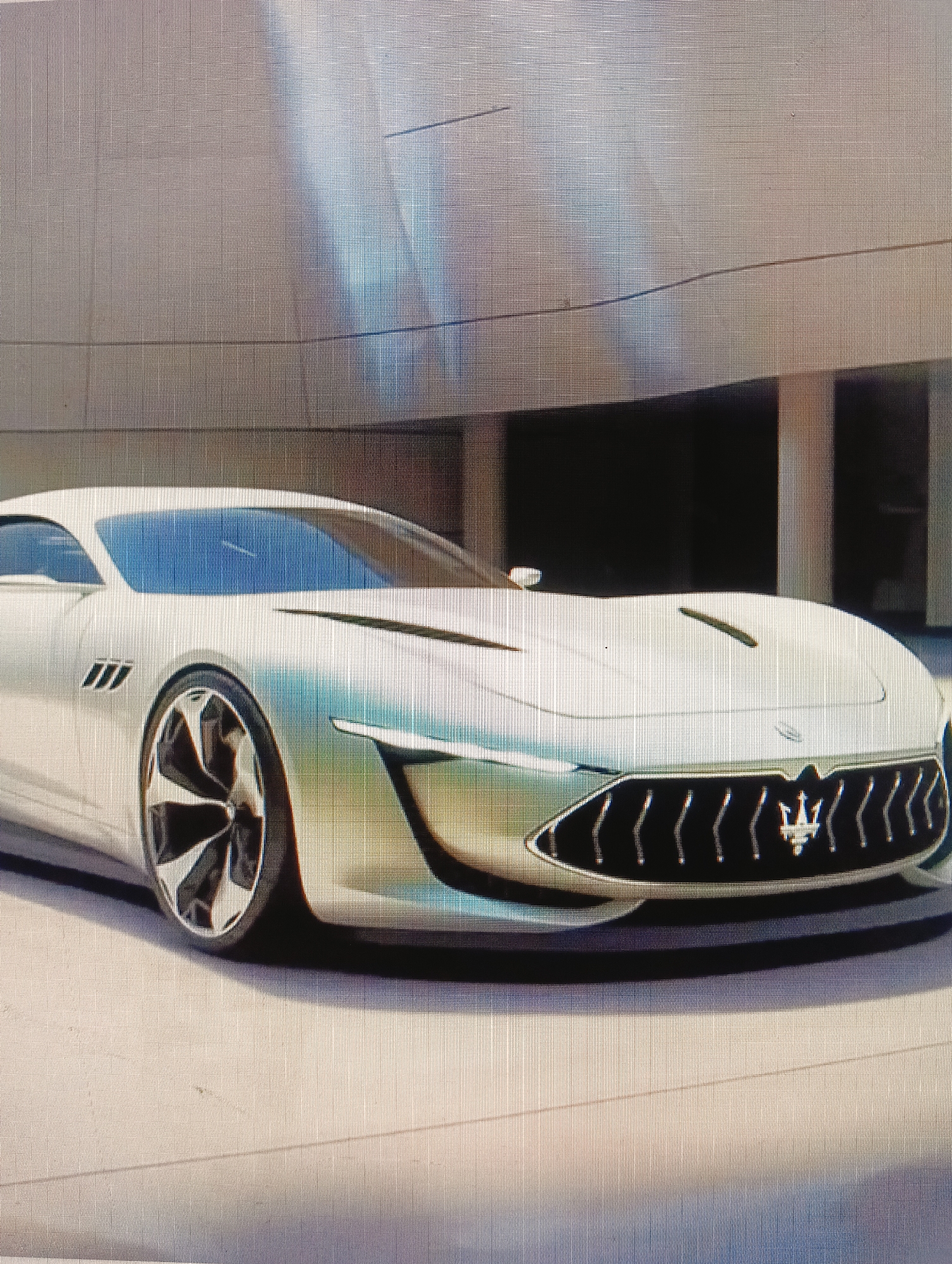 The image shows a futuristic concept car, a white coupe with a sleek design and a prominent grille. The car is positioned in what appears to be an outdoor urban setting, possibly a plaza or a street. The background features a modern building with a textured facade and dark openings, suggesting commercial or residential spaces. Sunlight casts shadows, indicating it's daytime. The car's design is characterized by sharp lines, a low profile, and large, intricate wheels. The front fascia has a distinctive grille with the Maserati trident logo. The overall aesthetic is one of luxury and advanced automotive engineering. The image does not contain any information that allows for identification of its specific location within the city of Sluck, Belarus, or any people or interactions.