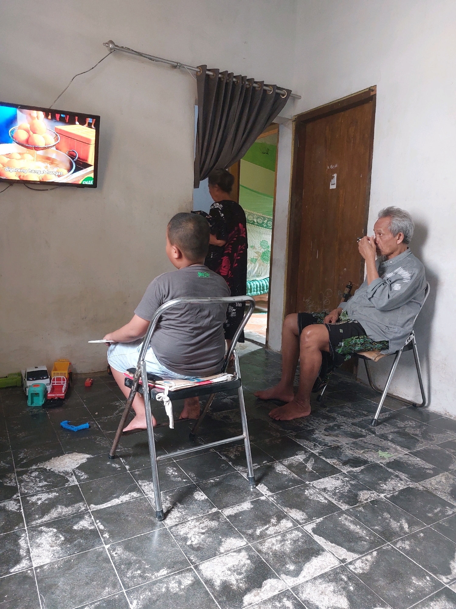In a sparsely furnished room, possibly in Sarangan, Indonesia, an older man with graying hair sits on a folding chair, smoking a cigarette. He wears a grey collared shirt and patterned shorts. To his left, a young boy sits on another folding chair, facing a television mounted on the wall. The television displays a cooking show featuring fried food. In the background, a woman stands near an open doorway, wearing a patterned dress. The room has tiled floors and bare walls, with curtains drawn over a window. Several toy trucks are scattered on the floor near the boy. The overall environment appears to be a modest living space.