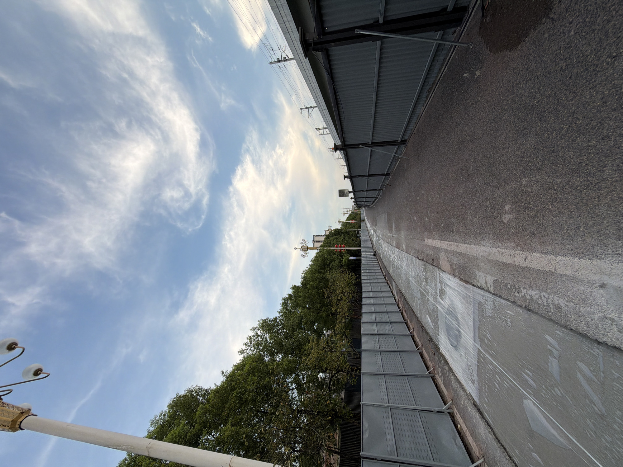 This image depicts a section of an elevated roadway or railway viaduct under construction or maintenance in Jiangjin District, Chongqing, China. The foreground shows a paved pathway, possibly a sidewalk or bike lane, with a section covered in wet concrete or a similar material, suggesting recent work. A grey, corrugated metal fence runs along the left side of the pathway, separating it from a treeline and what appears to be a body of water or a wet area. On the right side of the pathway, another grey metal fence of similar construction borders the roadway. Overhead, an elevated structure, likely the viaduct, is visible with its supporting columns and railway tracks. The sky is blue with scattered white clouds, indicating daytime. A streetlamp with multiple globes stands on the left side of the image, near the beginning of the pathway. The overall scene conveys an urban environment undergoing infrastructure development.
