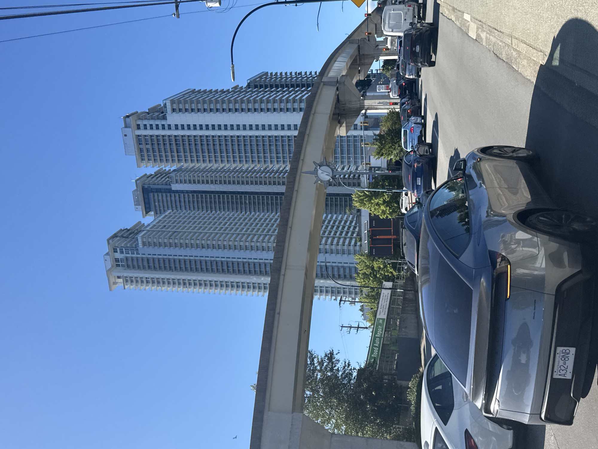 The image depicts a sunny day in Burnaby, Canada, with a view of multiple high-rise apartment buildings in the background. A monorail track is visible in the foreground, supported by concrete pillars. Several cars are on the road, indicating traffic congestion. One car in the foreground is a grey electric SUV, and a white sedan is to its left. The sky is clear blue, and trees are present on the sides of the road. A sign for "Road Auto Repair" is visible on a building.