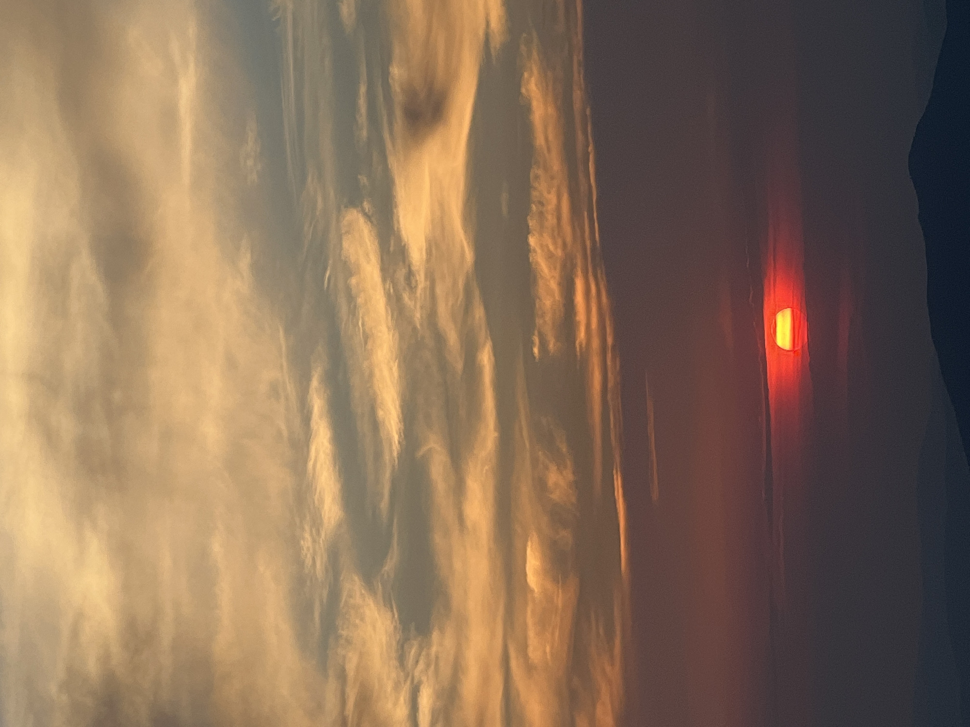 The image depicts a sunset over a mountainous landscape, likely in Luhy, Ukraine. The sky is filled with dramatic clouds, painted in hues of orange, yellow, and muted gray, indicative of the setting sun. The sun itself is a bright, vibrant red disk, partially obscured by a thin band of clouds near the horizon. Below the sky, dark, silhouetted mountains form a jagged line against the twilight. The overall impression is one of natural beauty and the serene transition from day to night. There are no people or man-made structures visible in the frame, focusing solely on the celestial event and the natural topography.