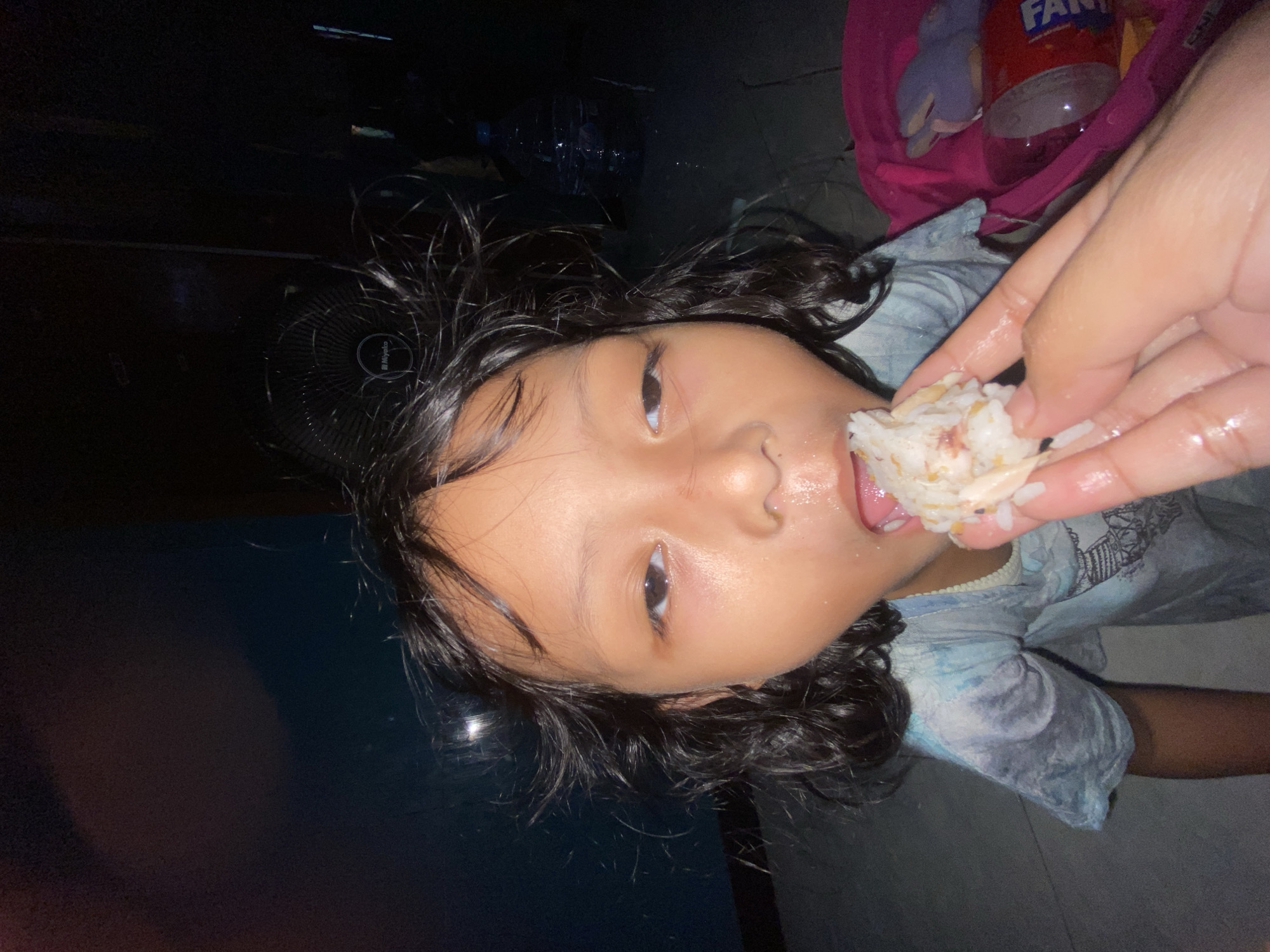 A young child with dark hair is being fed a bite of rice by a hand. The child's face is positioned centrally in the image, with their mouth open, ready to eat. The hand holding the rice appears in the upper right corner. A bag with a can of Fanta is visible nearby, along with other miscellaneous items. The background is dark. Based on the information provided, the location of the image is Gelam, Indonesia.