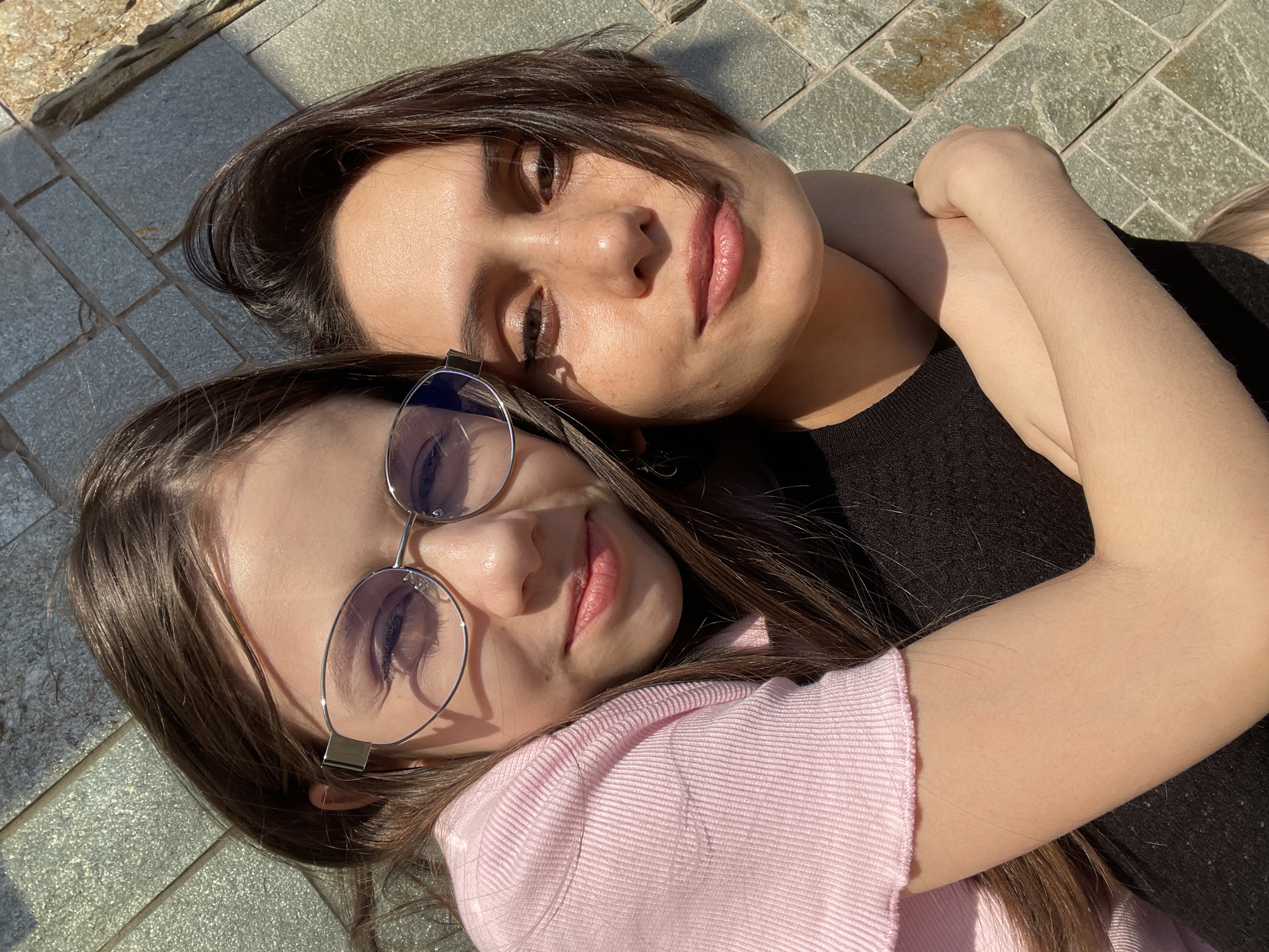 Two women are lying on a paved surface made of square gray tiles. The woman at the top of the frame has dark hair and is wearing a black top. Her arm is around the other woman. The woman at the bottom of the frame has brown hair, glasses, and is wearing a pink top. The location is likely Izhevsk, Russia based on the style of pavement.