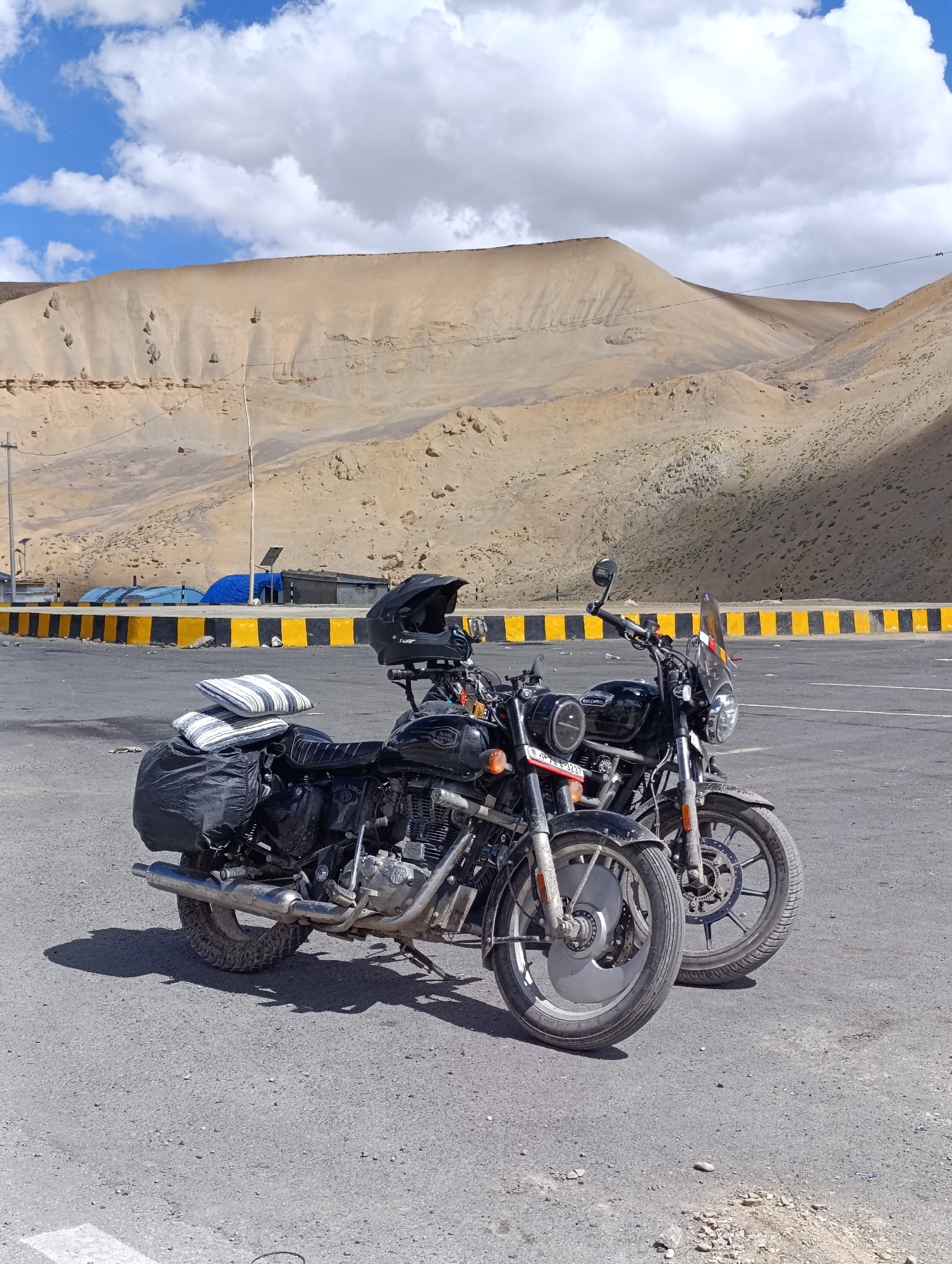 Two black Royal Enfield motorcycles are parked on an asphalt road in Pang, India. The motorcycle in the foreground is equipped with saddlebags and two striped pillows secured to the rear seat. A black helmet rests on the handlebars of this motorcycle. The second motorcycle, parked slightly behind and to the right, has a windshield. Both motorcycles have visible license plates and chrome accents.
In the background, a barren, sandy mountain landscape dominates the scene under a partly cloudy blue sky. The road is bordered by a barrier with alternating black and yellow stripes. Behind the barrier, several structures with blue and black tarps are visible, suggesting some form of temporary shelter or storage. Power lines and poles are present in the middle ground. The overall environment appears dry and arid, characteristic of a high-altitude desert region.