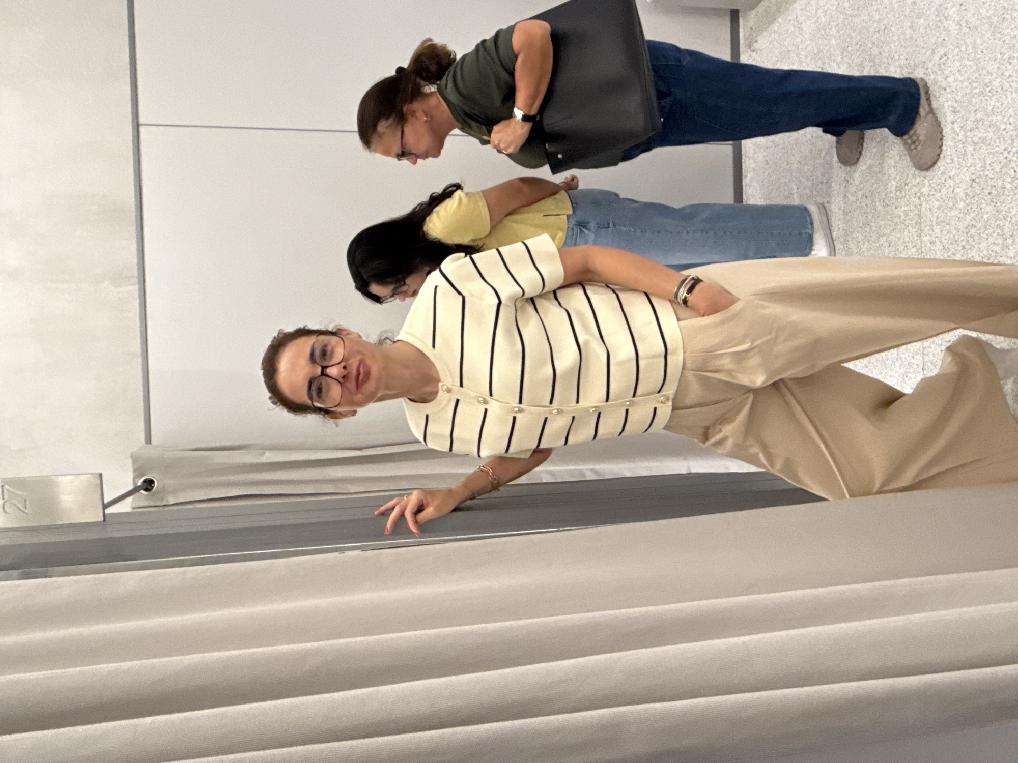 The image depicts three women in what appears to be a dressing room or fitting area, potentially in a retail store. The woman in the foreground is partially obscured by a privacy curtain; she is wearing glasses, a striped shirt with button details, and khaki-colored pants. Her hand is extended towards the curtain. Behind her are two other women. The first is wearing a yellow shirt and jeans. The second is wearing a dark green shirt and jeans and has a black bag. The background consists of a white wall and a light-colored speckled floor. The location, Matosinhos, Portugal, can not be determined from only visual data of the image.