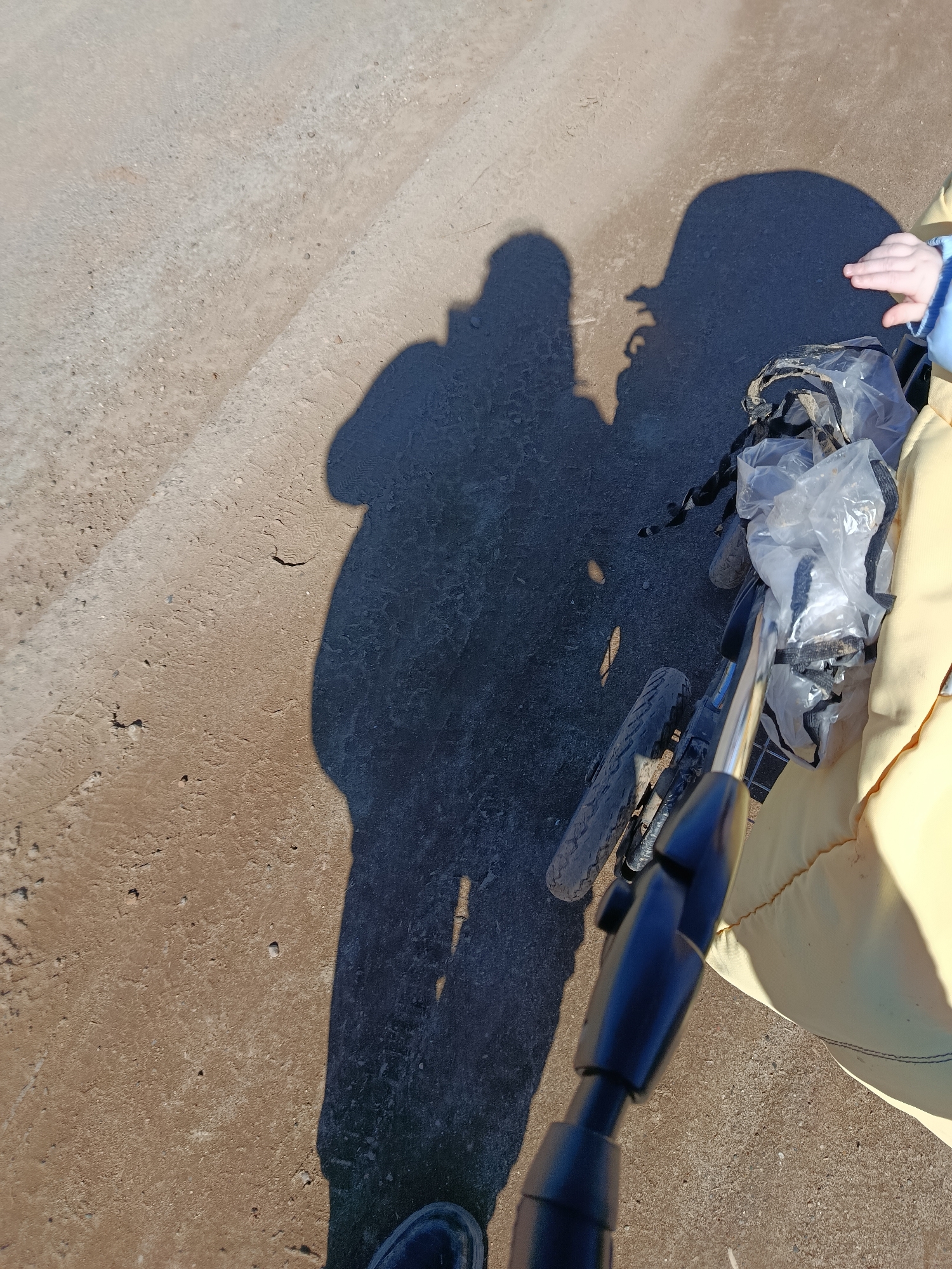 A person is pushing a stroller on a paved surface. The person's shadow is visible, along with the shadow of the stroller. A child's hand is visible in the stroller. The stroller has a plastic bag attached. One of the stroller's wheels is visible. The paved surface has a rough texture. The location is not discernible from the image.