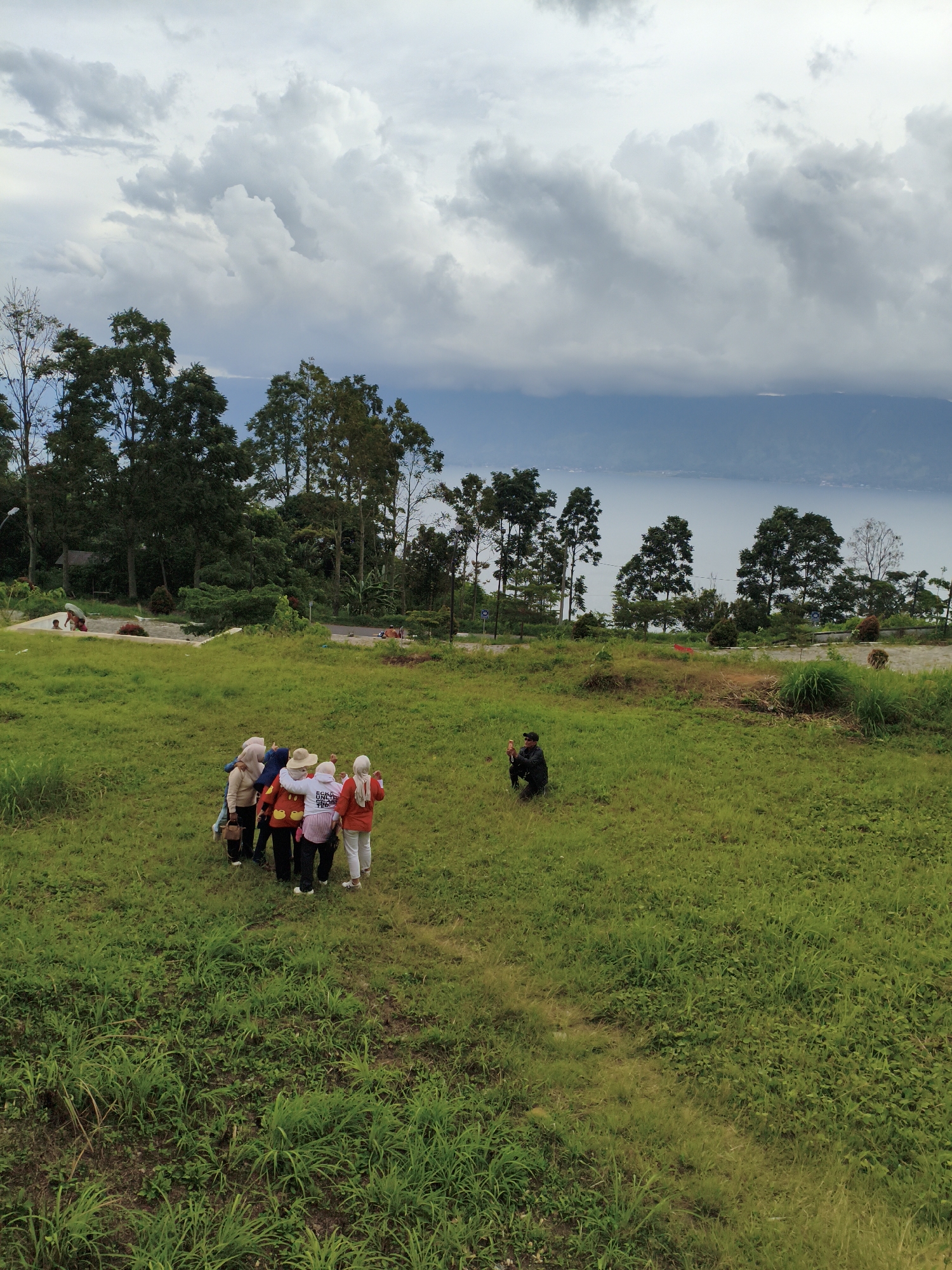 The image shows a group of five women standing close together in a grassy field, looking away from the camera. They are wearing headscarves and casual clothing. To the right of the group, a person is crouched down, appearing to take a photograph with a phone. In the background, there is a body of water, possibly a lake, bordered by a line of trees and hills. The sky is cloudy with dramatic cloud formations. The location is identified as a location in Pem.tambun Raya, Indonesia.