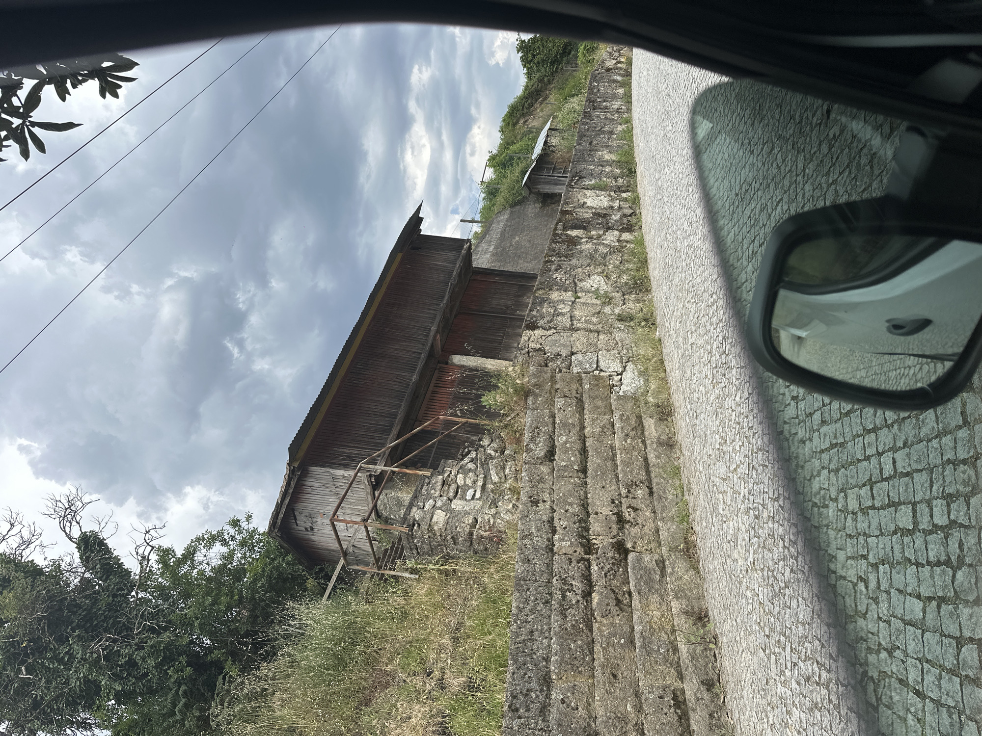 The image shows a view from inside a vehicle, capturing a street scene in Amarante, Portugal. A weathered wooden building with a rusty metal railing sits adjacent to a stone retaining wall with integrated steps. The stone wall runs alongside a narrow cobblestone street. Overcast skies are visible in the background. A side mirror of the vehicle is in the frame, reflecting the street. Vegetation grows around the base of the building and along the wall.