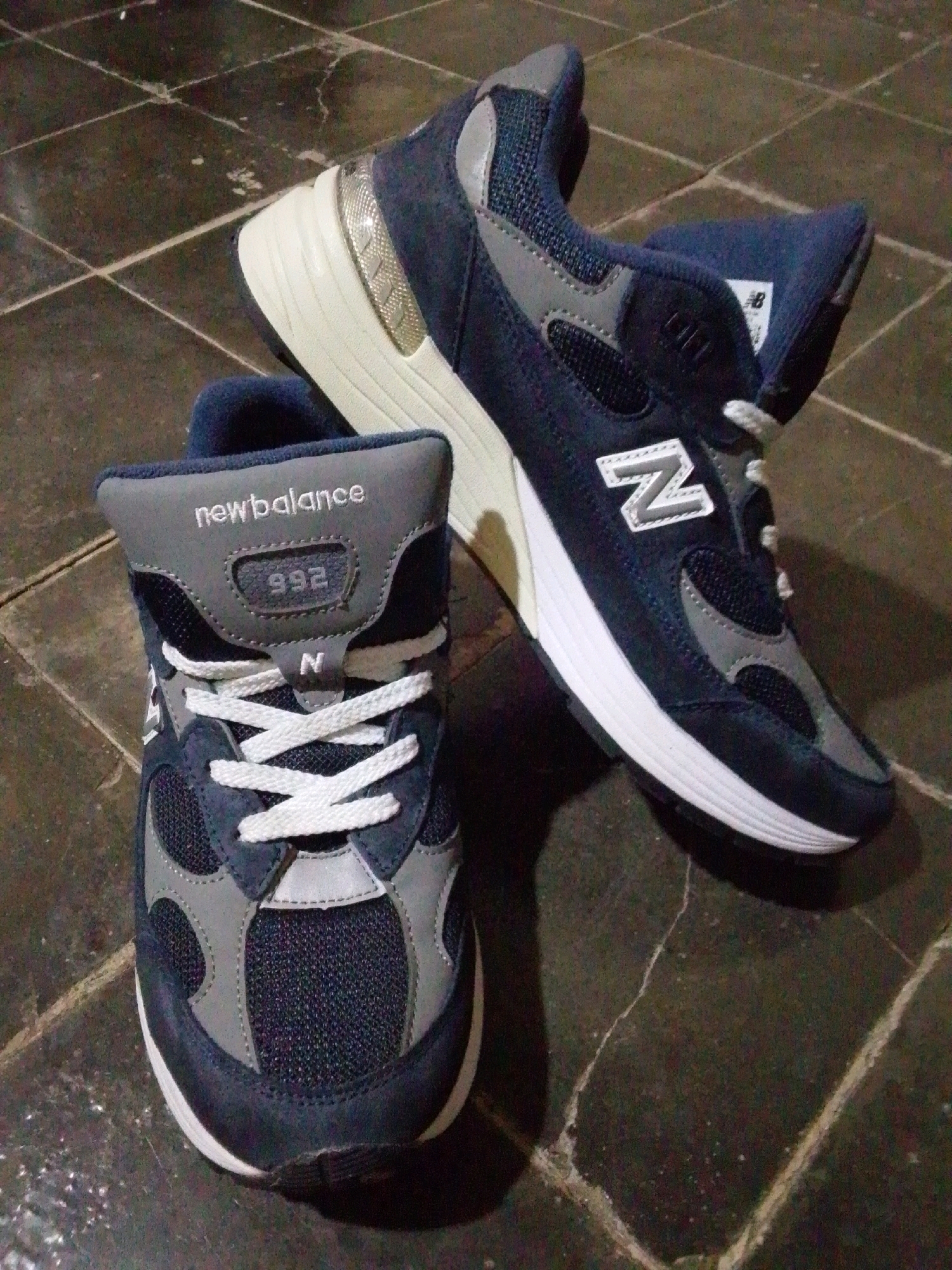 This is a forensic, matter-of-fact description of the provided image.
The image displays a pair of New Balance 992 sneakers placed on a dark, tiled floor. One shoe is positioned closer to the viewer, lying on its side with the sole facing upwards. The other shoe is placed behind and slightly above it, also on its side, with its sole facing away. Both shoes are primarily navy blue and grey, with white laces and accents. The New Balance logo is visible on both shoes, along with the model number "992" on the tongue of the shoe in the foreground. The tiled floor appears to be indoors, with visible grout lines and a slightly reflective surface. There are no people or any other objects depicted in the image. The lighting suggests an indoor environment with artificial light. The image does not provide any information about the city of Wirun, Indonesia, as it is not visible or referenced within the scene.