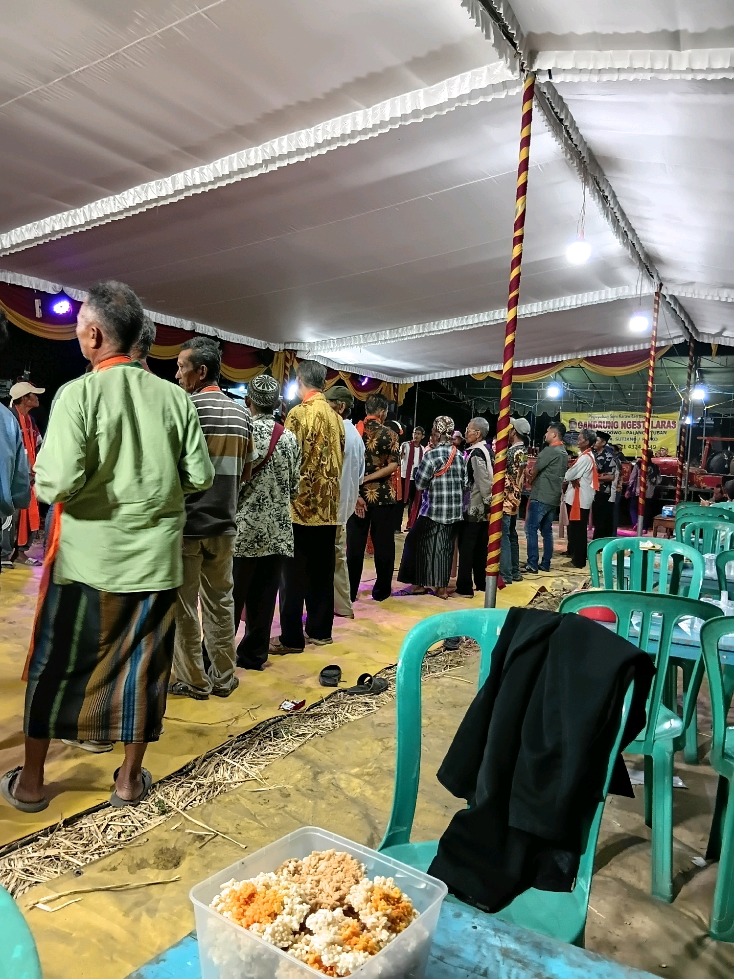 The image depicts an outdoor gathering under a large white tent, likely a community event in Soko, Indonesia. Several individuals, predominantly men, are standing and conversing. The attire suggests a formal or semi-formal occasion, with some men wearing traditional patterned shirts and sarongs. The ground is covered with a yellow tarp and straw. Green plastic chairs are visible, some with a black jacket draped over them. In the foreground, a clear plastic container holds a light-colored snack, possibly crackers or puffed rice, mixed with orange pieces. A blue table is in the immediate foreground. The tent is supported by red and yellow striped poles and illuminated by overhead lights. In the background, a banner with Indonesian text is partially visible, along with what appears to be a stage or performance area with musical instruments. The overall scene suggests a cultural event or celebration.