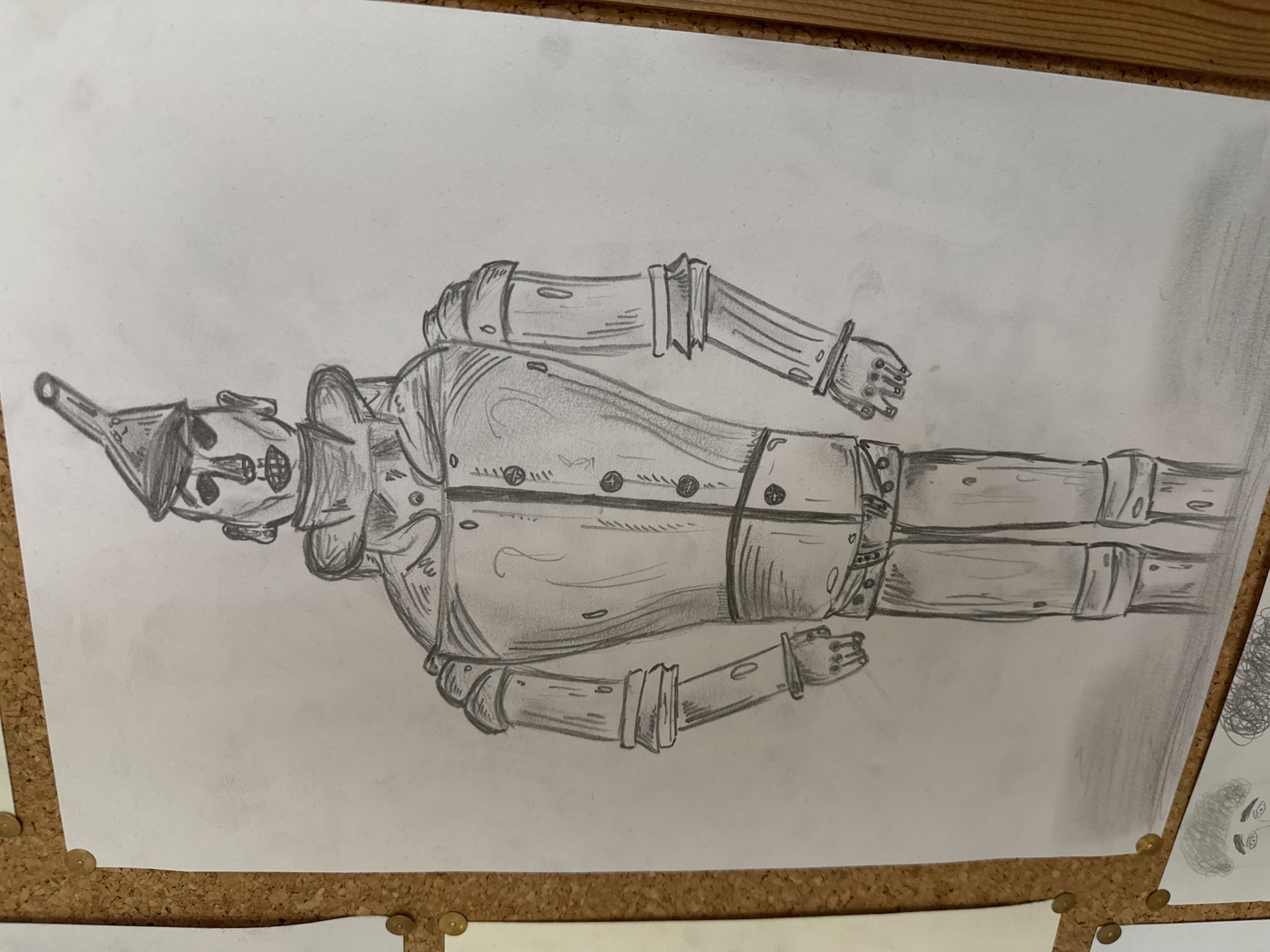 The media file features a detailed pencil drawing of a character resembling the Tin Woodman from "The Wizard of Oz." The full-body illustration depicts the figure standing upright, rendered with shading to suggest metallic surfaces and textures. Key features include a funnel-shaped hat, a riveted chest and limbs, articulated joints on the arms and legs, and stylized hands with hinged fingers. The character's face is simple, with vacant, circular eyes and a mouth resembling a hinged mechanism.
The drawing is presented on a sheet of white paper, which is secured to a cork board with several gold-colored thumbtacks visible along the top and bottom edges. The cork board suggests an indoor setting, possibly a studio or display area. No specific activity is depicted within the drawing, as it is a static portrait. There is no visible text in the image. The main image shows the drawing oriented horizontally, with the character lying on its side.