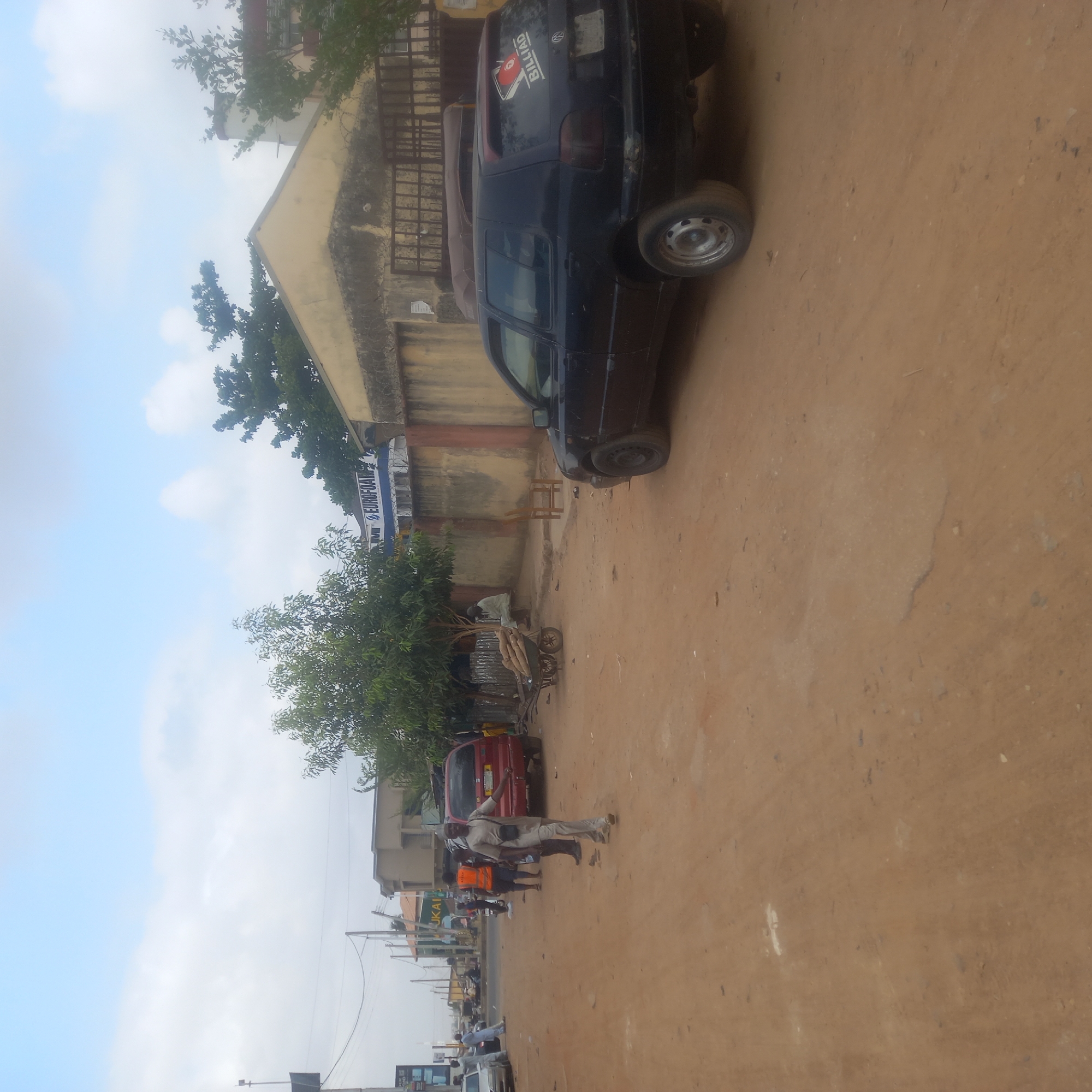 The image captures a lively street scene, likely in Dadi Riba, Nigeria, on an unpaved, reddish-brown dirt road. The sky above is partly cloudy, suggesting daytime.
Along the right side of the street, several buildings are visible, including one with a yellow facade and another featuring a blue and white sign displaying the text "EUROFOAM." Trees are interspersed along the roadside and in front of some buildings.
Multiple vehicles are present. A black car, identified as a Volkswagen by its logo and featuring a "BILLIAD" sticker with a pool ball graphic on its rear window, is parked prominently on the right. Further down, a red car is partially visible. Closer to the foreground, a person attends to a three-wheeled cart loaded with what appear to be long, thin logs or agricultural produce.
Several people are seen on the street. One man, dressed in a light-colored tunic and trousers, walks or stands towards the center of the frame. Another individual in an orange vest is visible further down the street, and other figures are indistinctly seen in the distance.
The overall scene depicts everyday activity on a bustling street, characterized by modest architecture, a dusty environment, and routine pedestrian and vehicle traffic. Utility wires crisscross the sky, adding to the urban landscape. Another blue sign with "JUKAL" is partially visible further down the street.