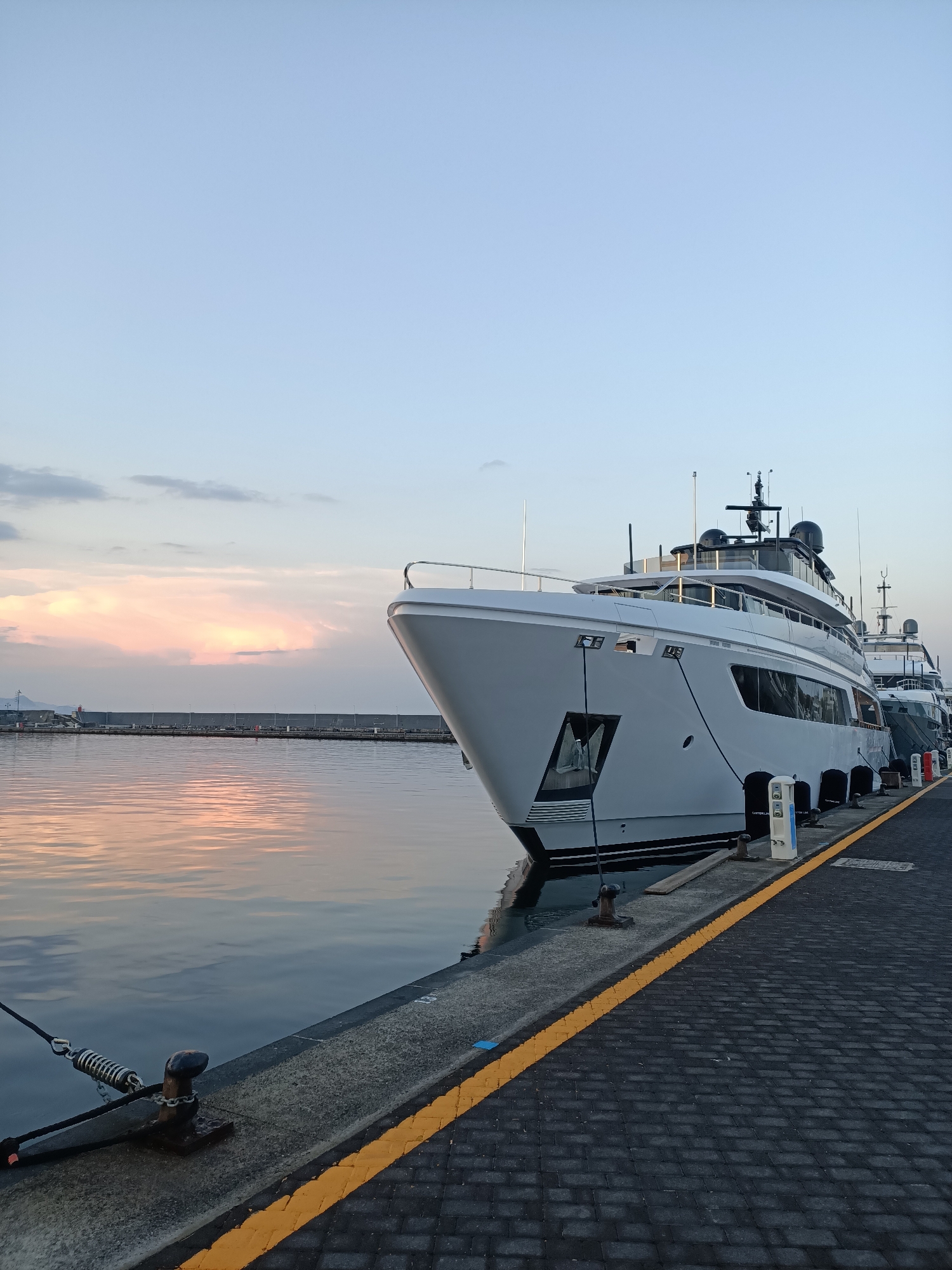 A wide shot captures a serene harbor scene at dawn or dusk, with the sky displaying a gentle gradient of pale blue to soft pink and orange hues, reflecting on the still water. In the foreground, a large, modern white yacht is prominently docked, its sleek design and polished surfaces gleaming. The yacht's bow faces the viewer, angled slightly towards the right side of the frame. The yacht is moored alongside a paved walkway lined with bollards. The walkway itself is constructed of dark paving stones, demarcated by a bright yellow line. Another yacht, partially visible, is docked behind the main subject. The background features a long, concrete breakwater or pier separating the harbor from the open sea, with a mountainous landscape faintly visible on the horizon. The overall atmosphere is one of tranquility and luxury.