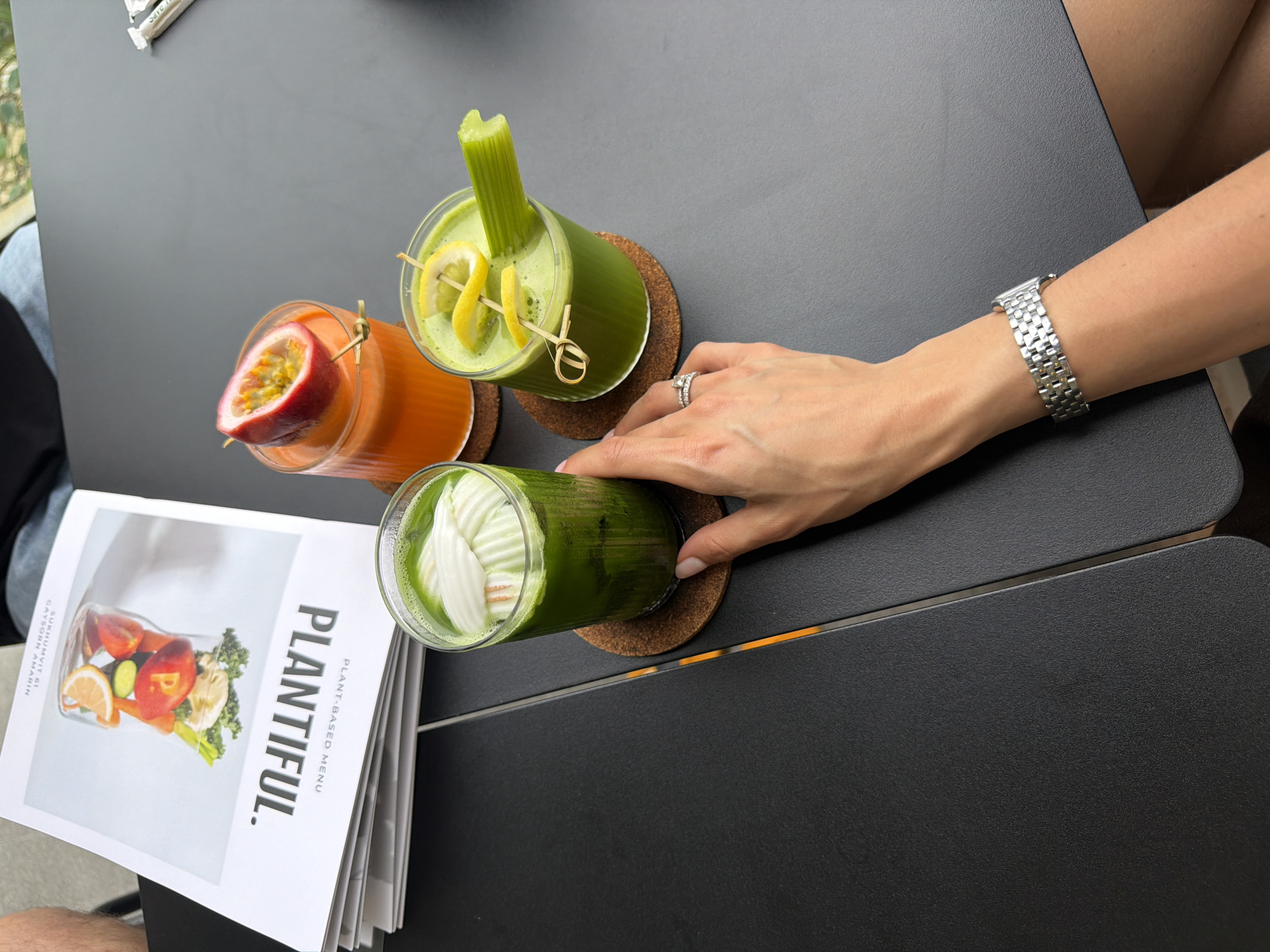 On a dark gray table, possibly outdoors at the Plantiful restaurant in Choeng Thale, Thailand, are three colorful drinks and a menu. The drinks, likely juices, are in clear glasses on round cork coasters. The first drink has an orange hue and is garnished with a half passion fruit. The second is green, featuring a celery stalk and lemon twists as garnish. The third drink, also green, has white garnishes. A woman’s hand, adorned with a silver watch and a diamond ring, rests near one of the drinks. A white menu with the word "Plantiful" is partially visible.