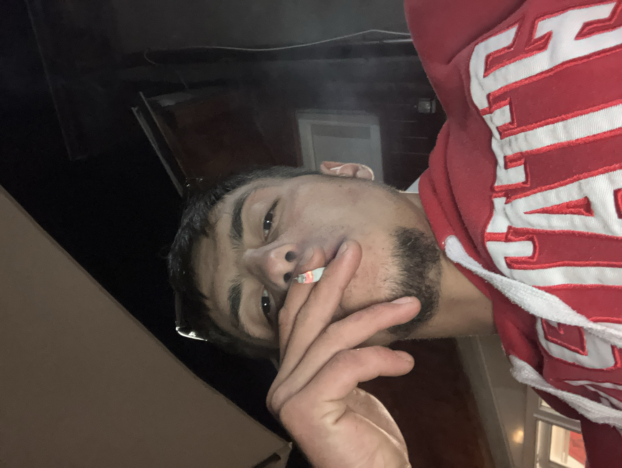 A young man is smoking a cigarette. He is wearing a red hooded sweatshirt with white lettering. The background is dark and appears to be outdoors at night. There are visible structures in the background, including what appears to be a wall and a window frame. The location is not discernible from the image.