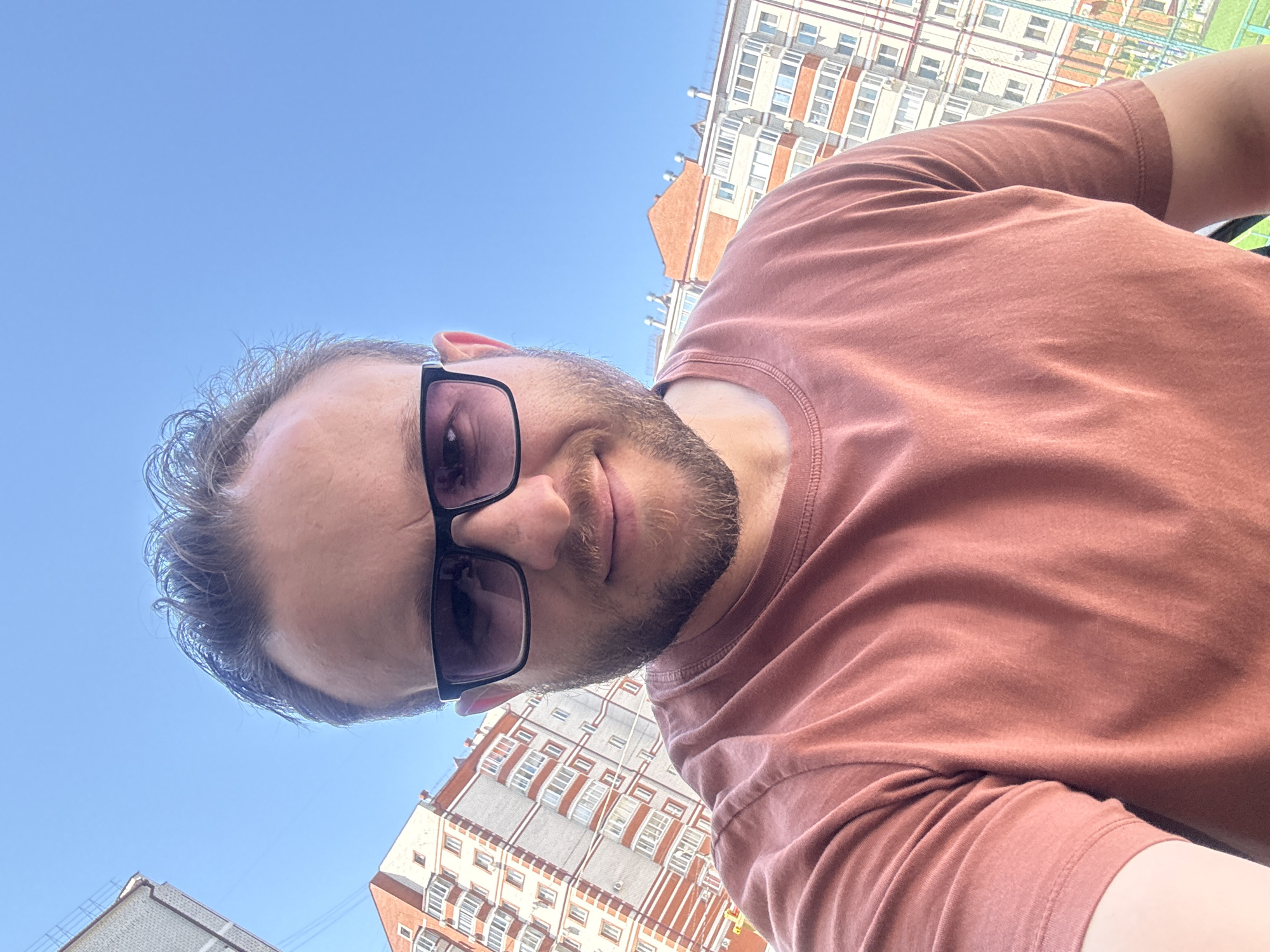 A man with short brown hair and a beard is taking a selfie outdoors. He is wearing dark sunglasses and a rust-colored t-shirt. Behind him, a modern apartment building with red and white brickwork is visible under a clear blue sky. The building appears to be in a city setting, and the context provided suggests the location is Chigiri, Russia.