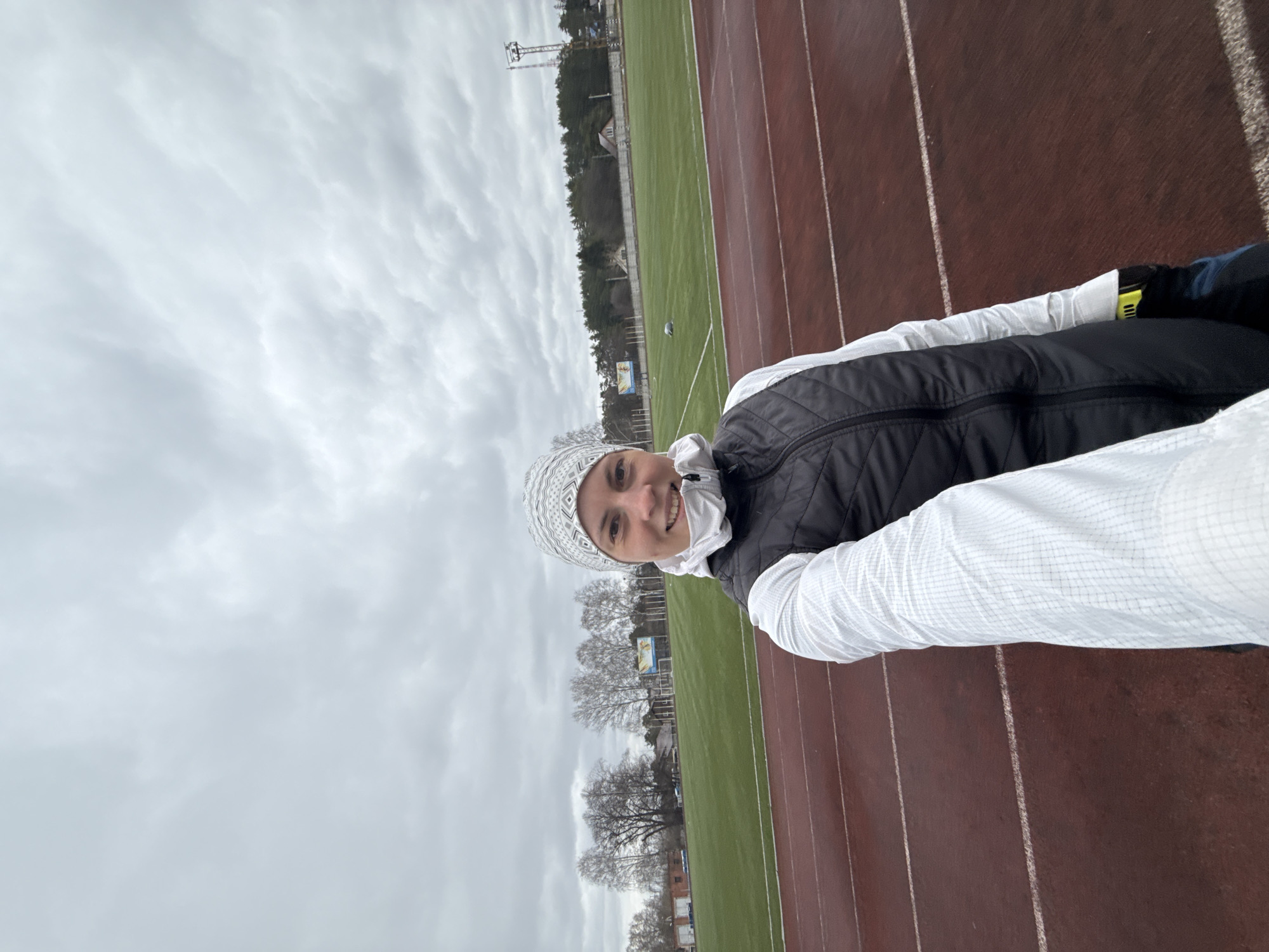 The image depicts a woman taking a selfie at an outdoor athletic stadium in Chaikovsky, Russia. The woman, positioned on the brown running track, is smiling pleasantly at the camera. She is dressed for cool weather, wearing a white patterned knitted hat, a white long-sleeved top, and a black quilted puffer vest. A small yellow patch is visible on her left arm, though any text on it is illegible.
The background features the stadium's facilities, including several lanes of a reddish-brown athletic track with faded white marking lines. Adjacent to the track is a large, vibrant green field, likely a football or soccer pitch. Beyond the field, a line of trees with bare branches suggests the season is either late autumn, winter, or early spring. In the distance, parts of stadium structures, including a tall light tower and some buildings, can be seen. The sky is uniformly overcast with a blanket of light grey clouds, indicating a cloudy daytime environment with subdued lighting. No discernible text is visible on any banners or signs in the background.