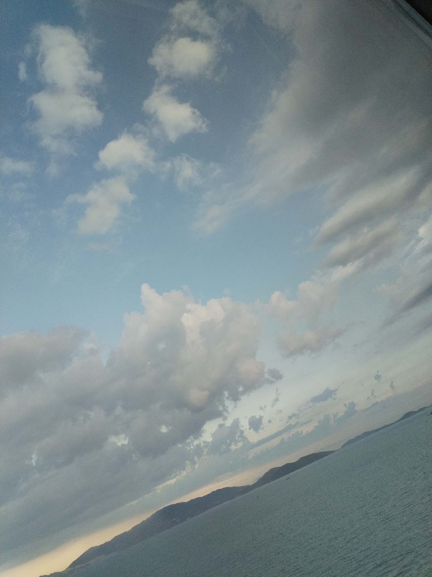 The image shows a view of Nha Trang Bay, Vietnam. The horizon line features landmasses, potentially islands or a distant coastline. The sea occupies the lower portion of the frame. The sky is filled with cumulus and stratus clouds, indicating varying weather conditions. The lighting suggests either early morning or late afternoon. No people, subjects, or actions are directly visible in the frame. The image composition is tilted, adding a dynamic perspective.