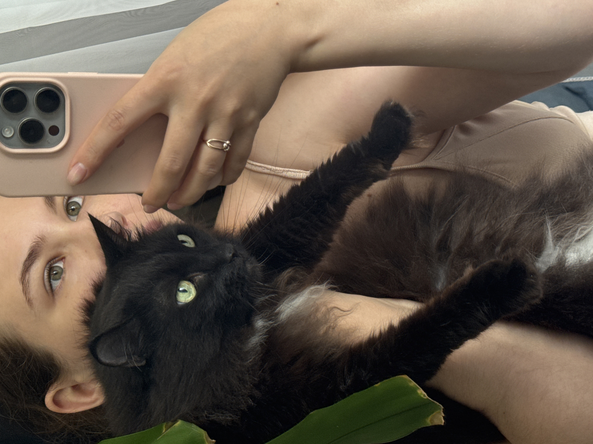 A young woman with light brown hair is taking a selfie with her black cat. She is holding the cat in her arms and looking at the phone's screen. The cat has green eyes and a white patch of fur on its chest. The woman is wearing a beige tank top and a gold ring on her right hand. The background is blurred and appears to be a room with a window and curtains. The scene is set in Chaikovsky, Russia.