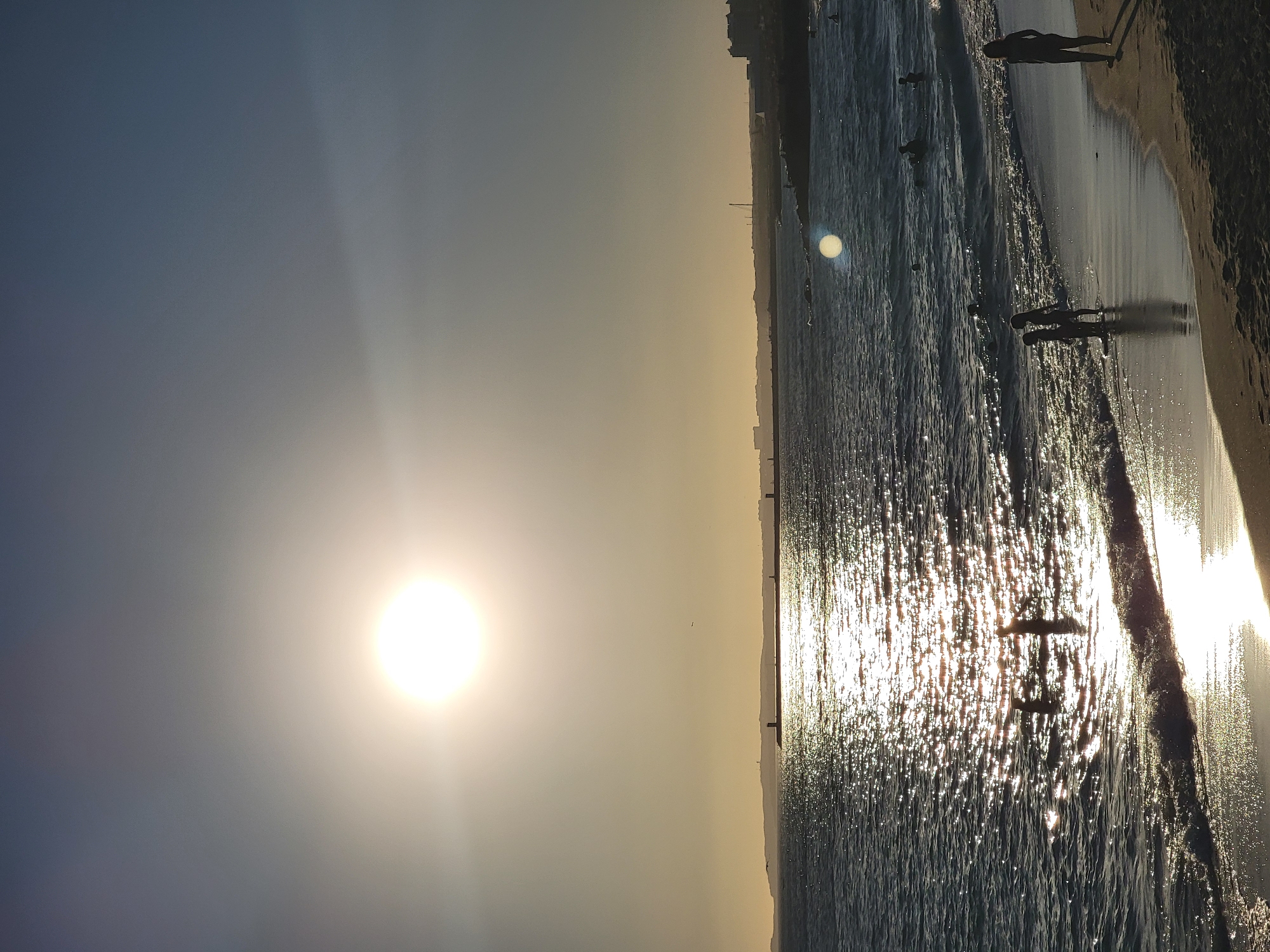 A vertical, eye-level shot captures a serene beach scene in Quarteira, Portugal, at sunset. The sun, a bright, glowing orb, is positioned in the upper left quadrant of the frame, casting a shimmering path across the choppy, dark blue ocean. The water reflects the sunlight, creating a dazzling, glittery surface.
A long, dark landmass stretches across the horizon, silhouetted against the hazy, golden sky. Several lighthouse-like structures are visible along this distant shore, hinting at a port or coastal defense. The sky transitions from a pale yellow near the horizon to a deeper blue towards the top of the image.
In the foreground, a sandy beach meets the water's edge. Three figures stand partially submerged in the shallow water near the left side of the frame, their forms silhouetted against the bright reflections. Further to the right, another lone figure stands on the wet sand, facing away from the camera, their shadow stretching across the beach. A few other indistinct figures can be seen further out in the water. The overall impression is one of a peaceful evening at the seaside, with the sun's descent creating a warm and inviting atmosphere.