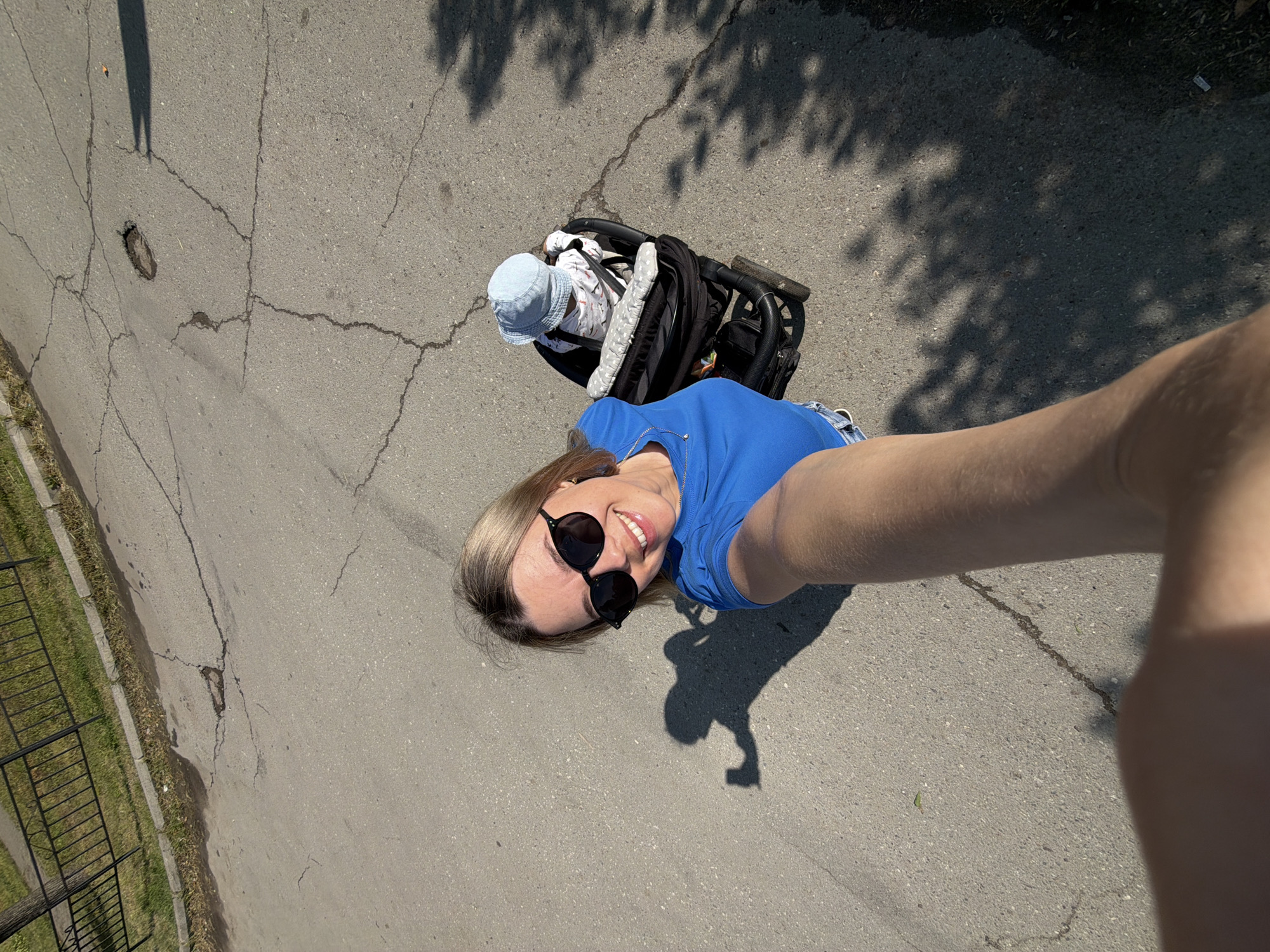 A woman is taking a selfie outdoors, holding her phone with her right arm extended. She is wearing a blue t-shirt and sunglasses, and is smiling. Beside her is a black stroller with a baby wearing a light blue hat. The scene takes place on an asphalt surface with cracks. A black metal fence and grass are visible in the upper left corner of the image. The sun casts shadows on the ground. The context of Chaikovsky, Russia is provided as the location.