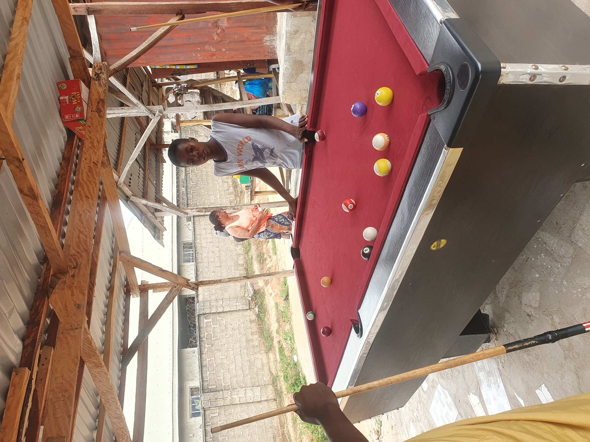 This image depicts an outdoor billiard setup in Lokoja, Nigeria. A young Black male, wearing a sleeveless white t-shirt with graphics, stands behind a red-felt pool table, seemingly preparing to play. His hands are resting on the table edge, and a pool cue is held by another person's hand in the foreground, positioned to the left. Several billiard balls are scattered across the table, including a black 8-ball, a white cue ball, a red and white striped ball (possibly a 1-ball), a solid yellow ball (possibly a 3-ball), a solid yellow ball (possibly a 7-ball), a solid red ball (possibly a 5-ball), a solid purple ball, and a green ball. In the background, a woman in an orange top and patterned skirt is visible, along with other indistinct figures and structures, suggesting a communal outdoor area. The setting includes a partially constructed wooden roof structure supported by beams, with corrugated metal roofing. A brick wall with barbed wire fencing is visible in the mid-ground, and a dirt and paved ground surface surrounds the pool table. The overall impression is a casual, open-air recreational space.