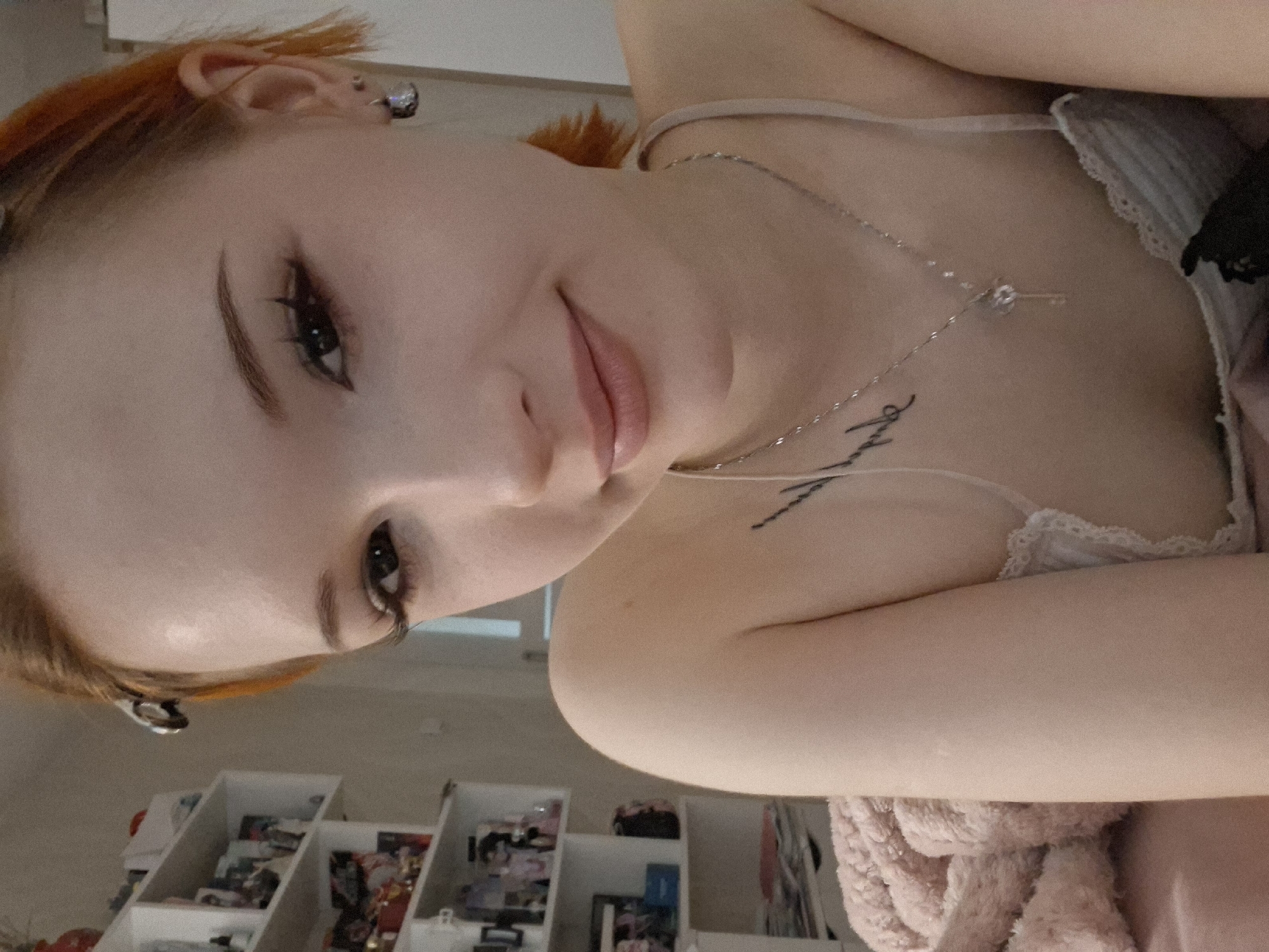 A young woman with fair skin and orange hair is pictured in a close-up, slightly rotated view. She has dark eyes and wears eyeliner. A silver necklace with a pendant is visible around her neck, along with a tattoo on her chest. She is wearing a light-colored top with lace detailing. In the background, out of focus, is a white shelf unit filled with various objects, including books and decorations. Based on the reference to Chaikovsky, Russia, the photo was likely taken in a residential environment in that city. No specific street or building can be identified.