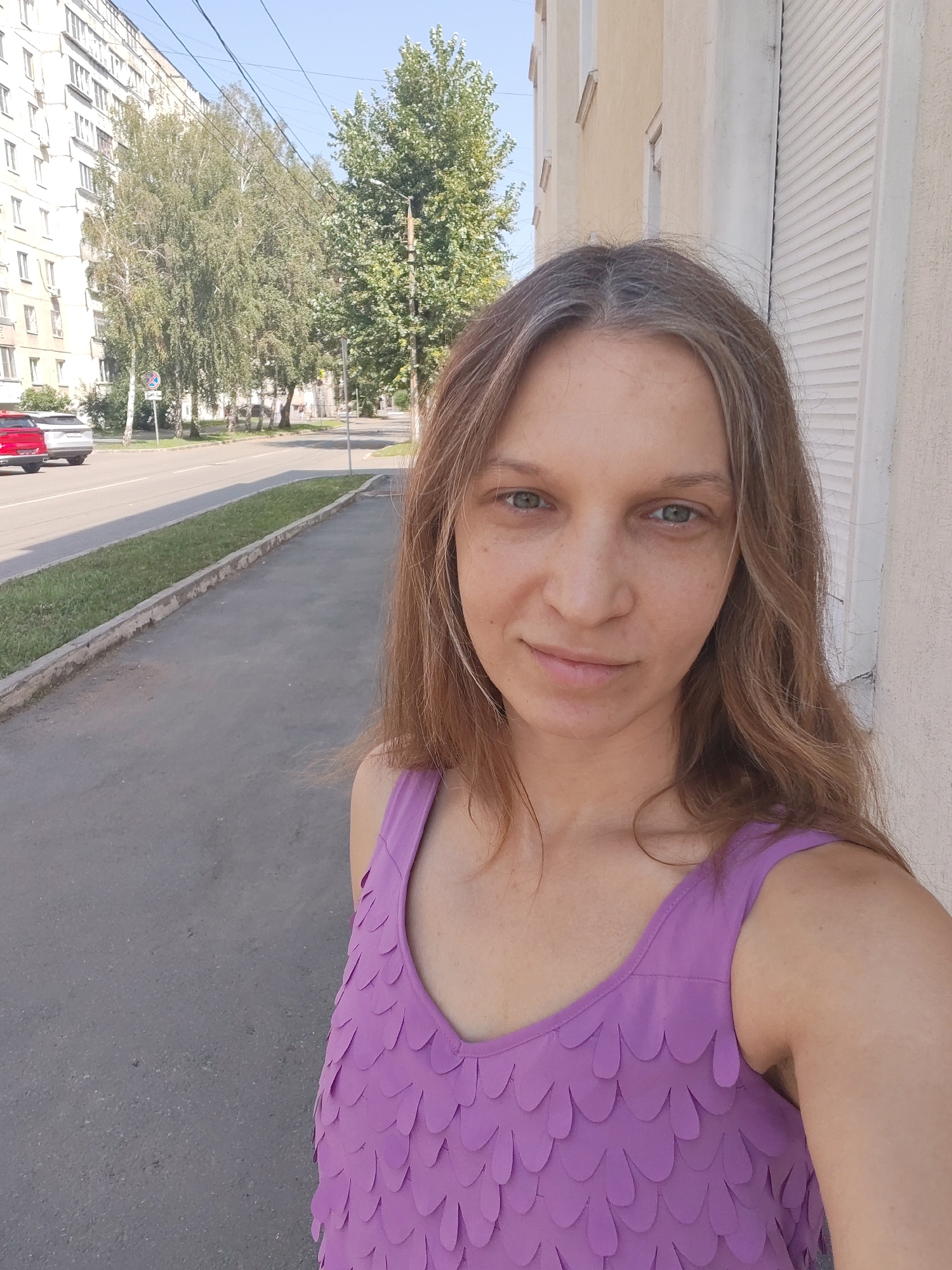 A woman is taking a selfie outdoors on a street. She has long, wavy brown hair with some graying and light blue eyes. She is wearing a purple tank top with a scalloped pattern. Behind her, there is a sidewalk and a street with a couple of cars. A multi-story apartment building and several trees are visible on the left side of the image. The building on the right has a shuttered window. The sky is clear and blue.