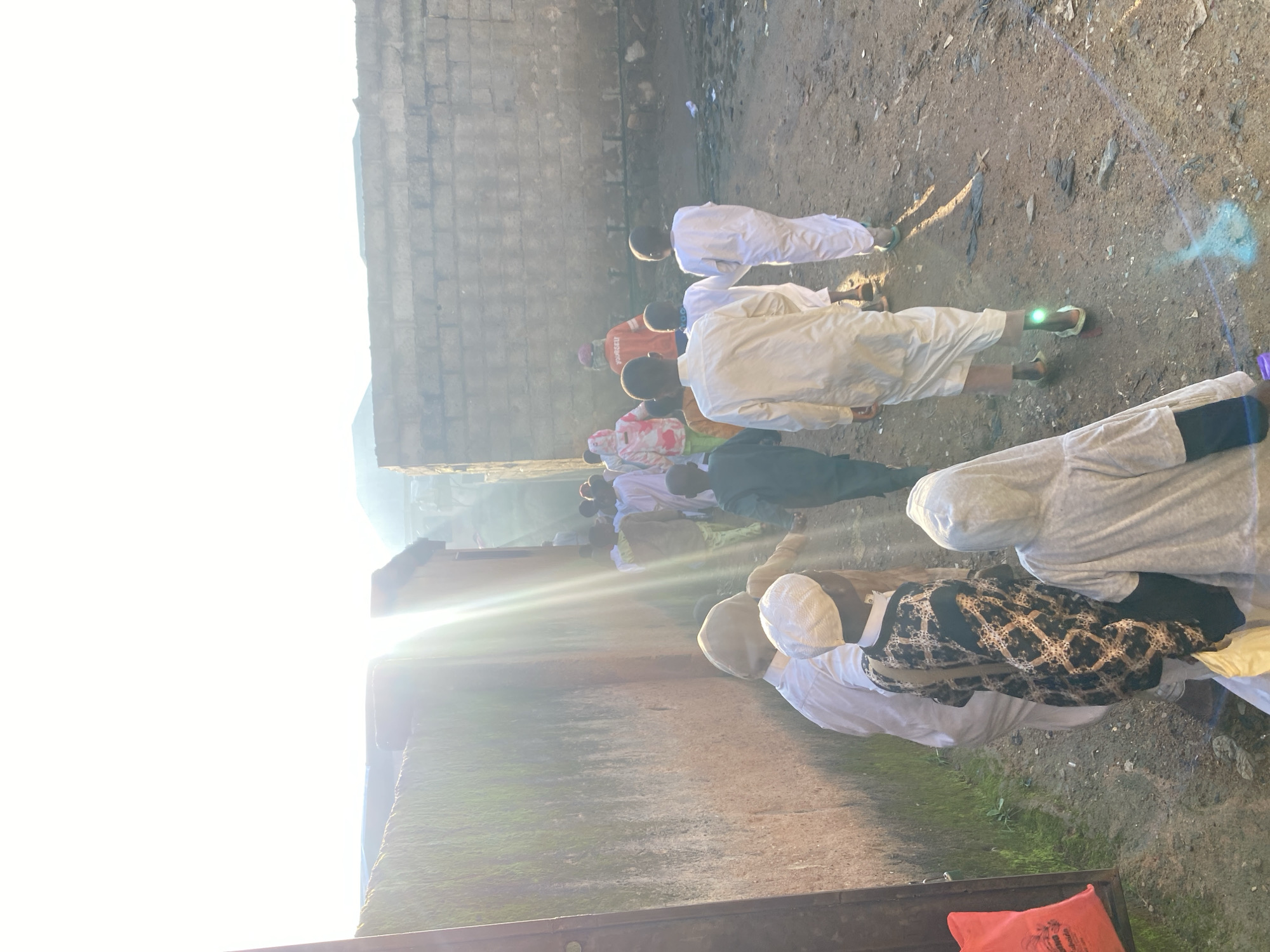 A group of children are walking away from the camera down a dirt path between two buildings. The building on the left is a reddish-brown color with moss on the lower portion of the wall. The building on the right is made of brick. Sunlight streams from the left side of the image, creating a lens flare that obstructs some of the details. The children are dressed in traditional attire, with many wearing white robes and some wearing patterned garments. One child in the foreground wears a grey hooded robe, and another in front of them wears a white skullcap and a patterned black and gold robe. A person wearing an orange vest with "YOUNGEU" visible on the back is walking with the children. The ground is dusty with some debris scattered about. The overall scene suggests a procession or movement of a group of young people in an outdoor urban environment in Jos, Nigeria.