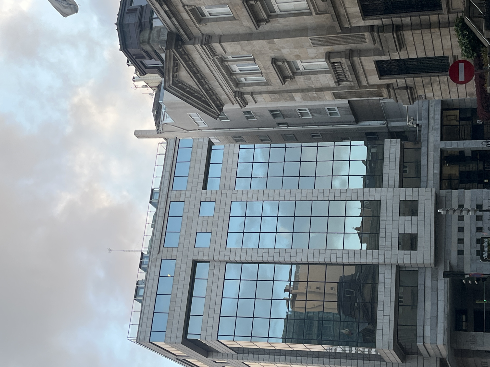 The image depicts a view looking up at buildings in the city of A Coruña, Spain. The main focus is on a modern building with a glass curtain wall reflecting the sky and surrounding buildings. Adjacent to the modern building is an older, more ornate building with traditional architectural details. A "Do Not Enter" sign is also visible.